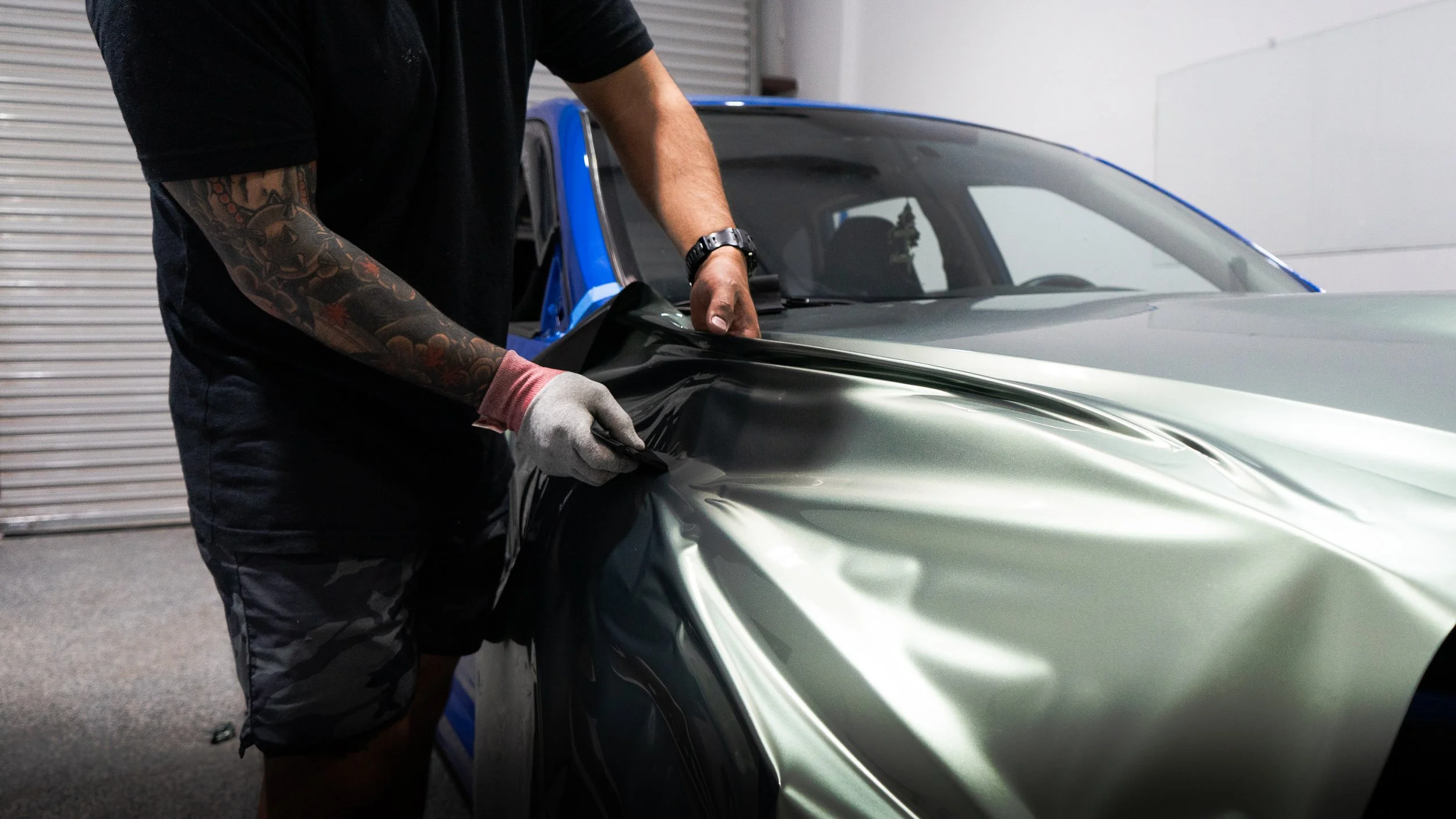 Person applying a wrap or vinyl decal to a metallic gray car in a garage.