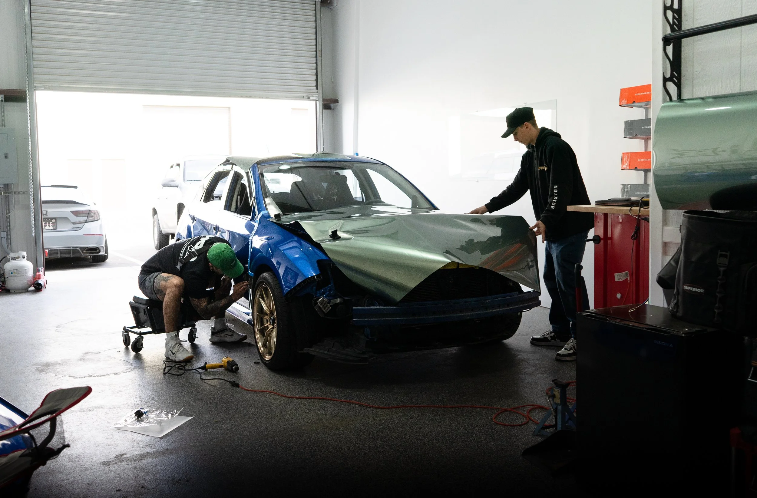 Two mechanics working on a race car inside a garage, one inspecting the front left wheel and the other handling the hood, with tools and equipment around.