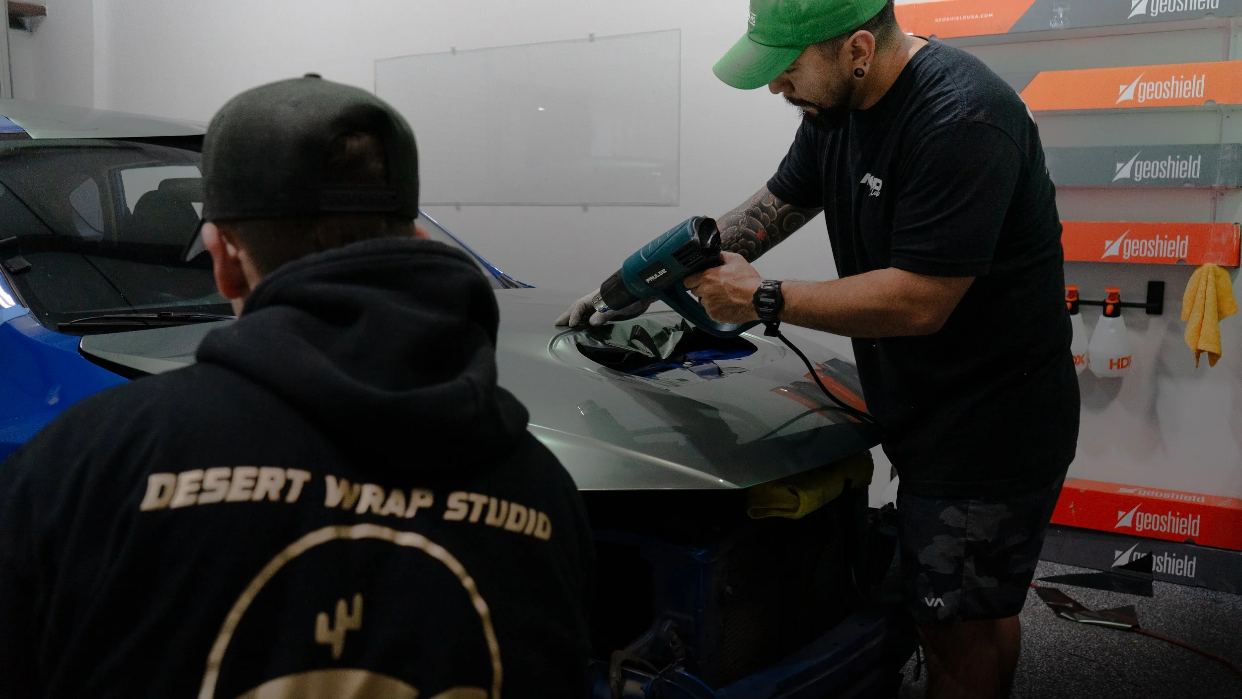 Two men working on a car in a garage, one using a heat gun to apply a black film, the other with his back to the camera wearing a hoodie that says 'Desert Wrap Studio'.