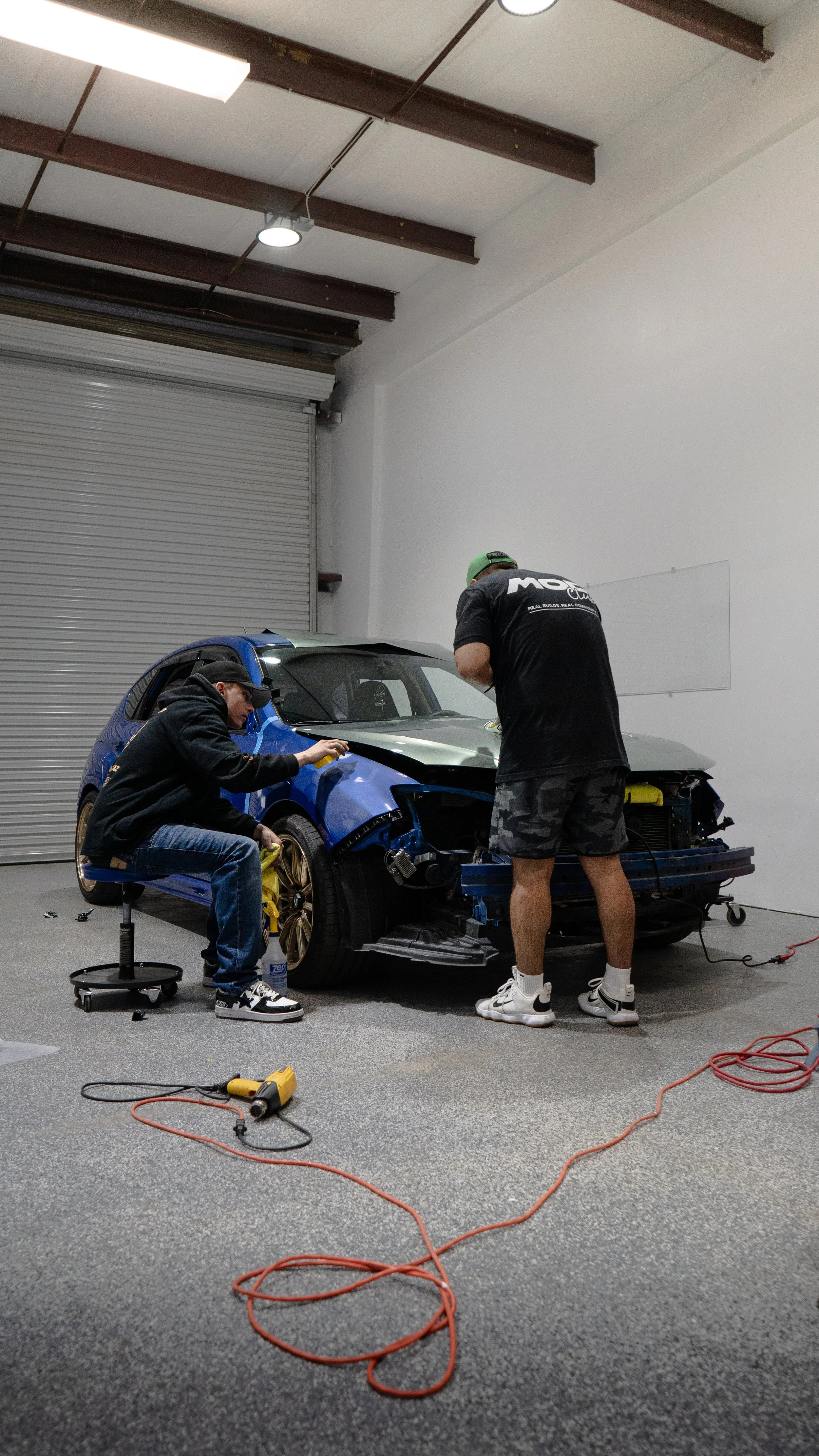 Two men working on a damaged race car inside a garage, one sitting and carefully working on the front wheel, the other standing and working on the front of the car. The car has a blue and black body with a silver hood, and the garage has a white wall and a large roll-up door in the background.