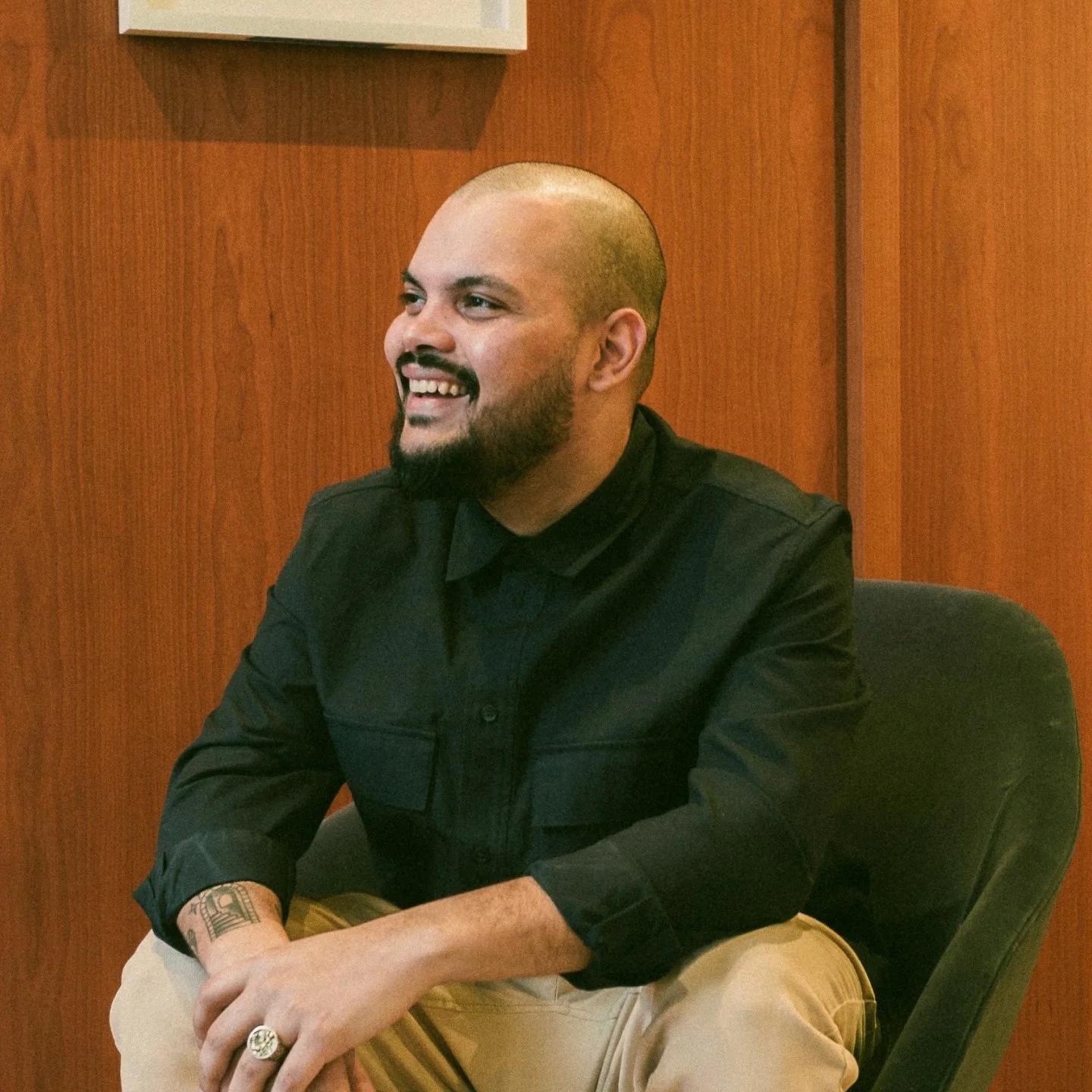 A smiling man with a beard and shaved head, wearing a black shirt and khaki pants, sitting in a black chair against a wooden-paneled wall.
