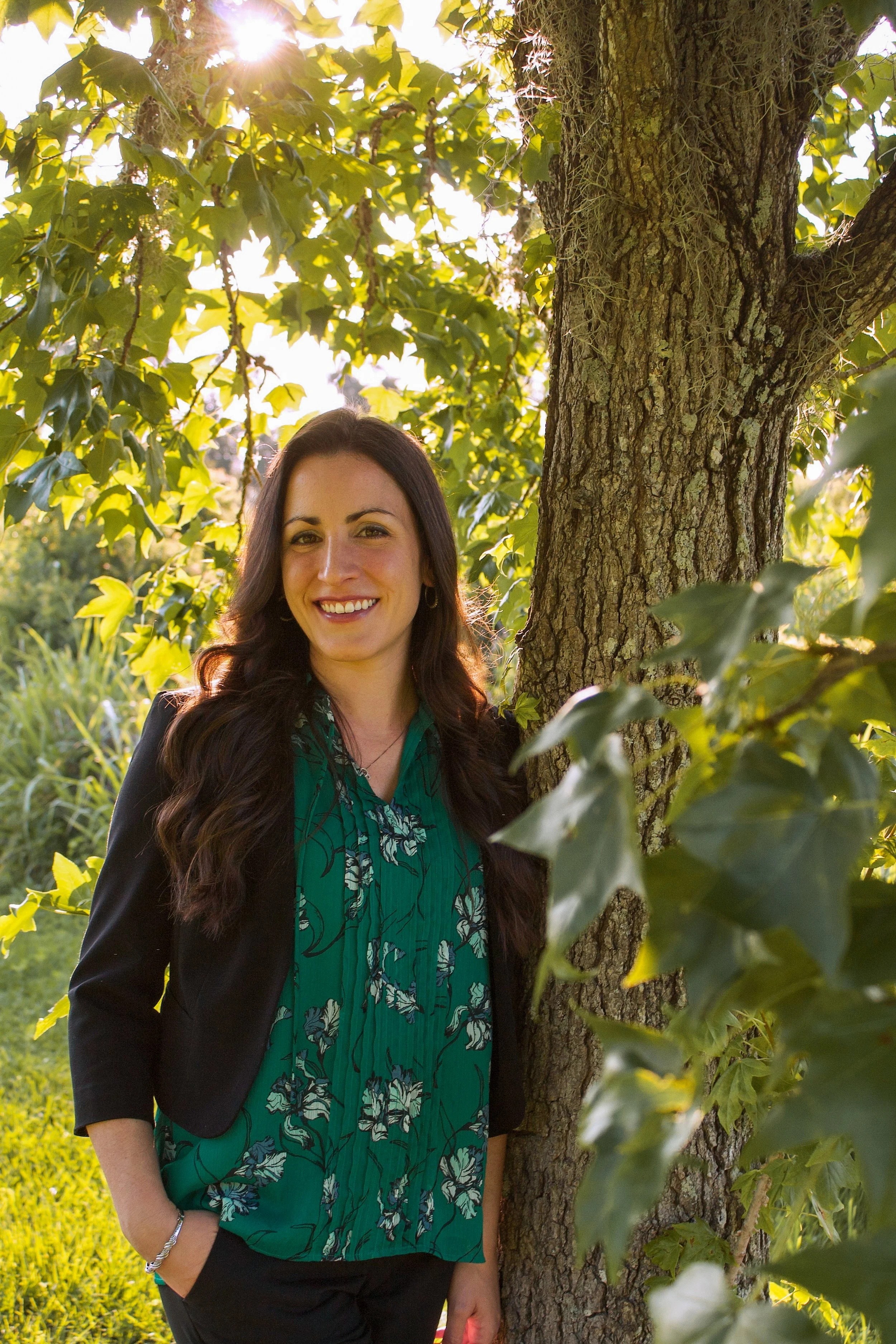 A woman with long dark hair smiling outdoors beside a tree with green leaves sunlight shining through.