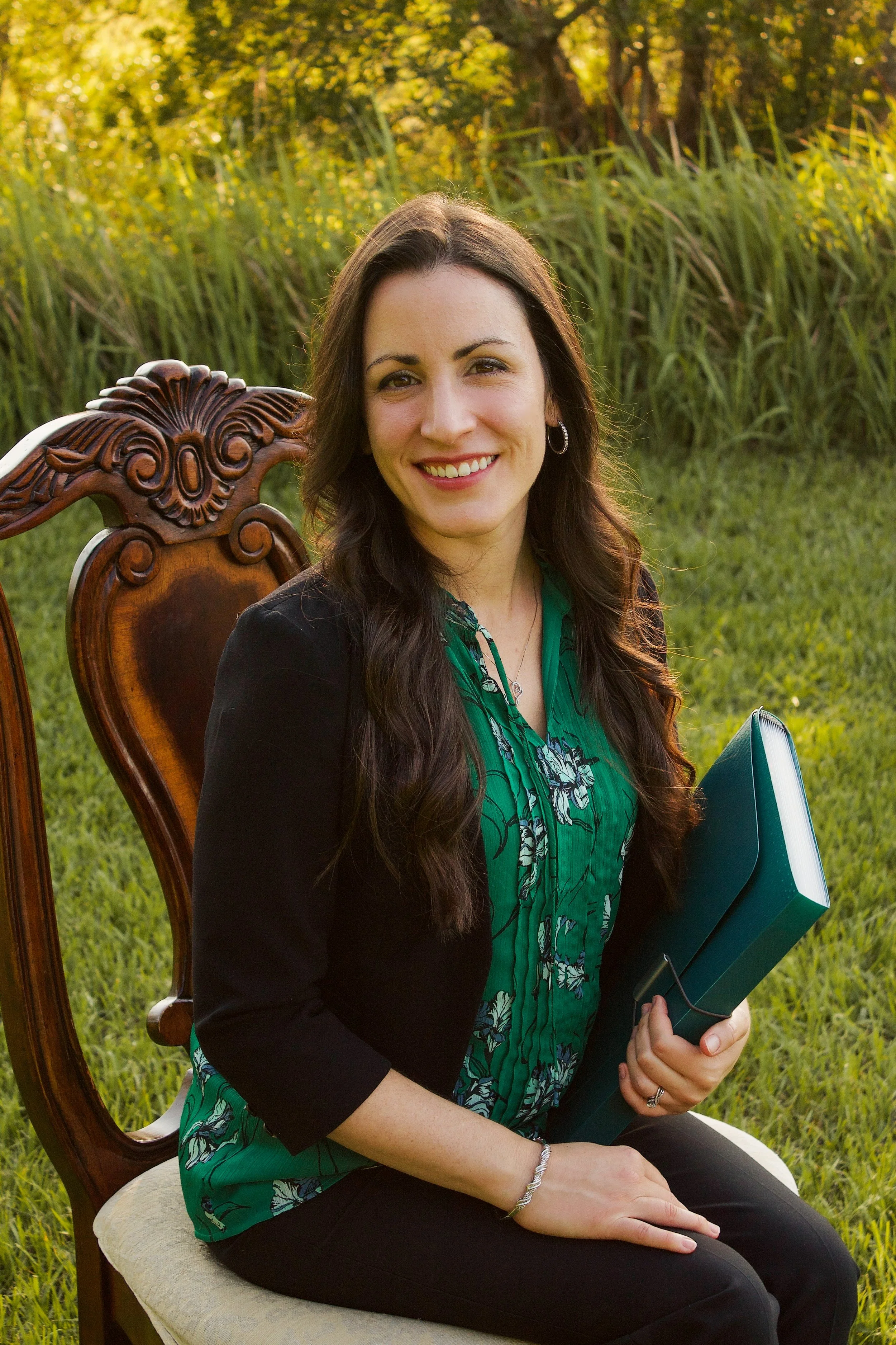 A woman with long wavy brown hair, smiling, sitting on a wooden chair outdoors, holding a blue binder, with green grass and trees in the background during daylight.