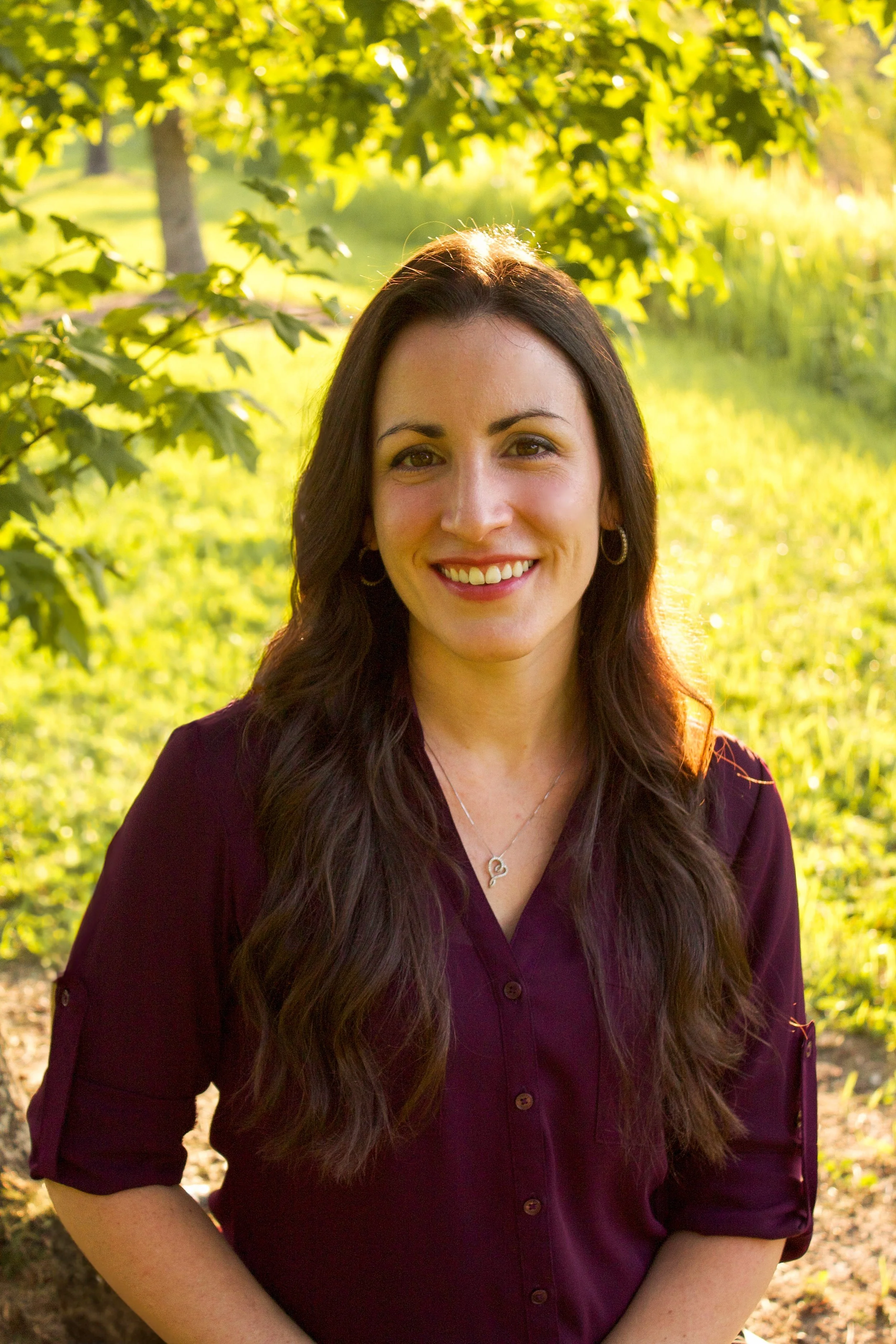 A woman with long dark brown hair and hoop earrings smiling outdoors in a park on a sunny day.