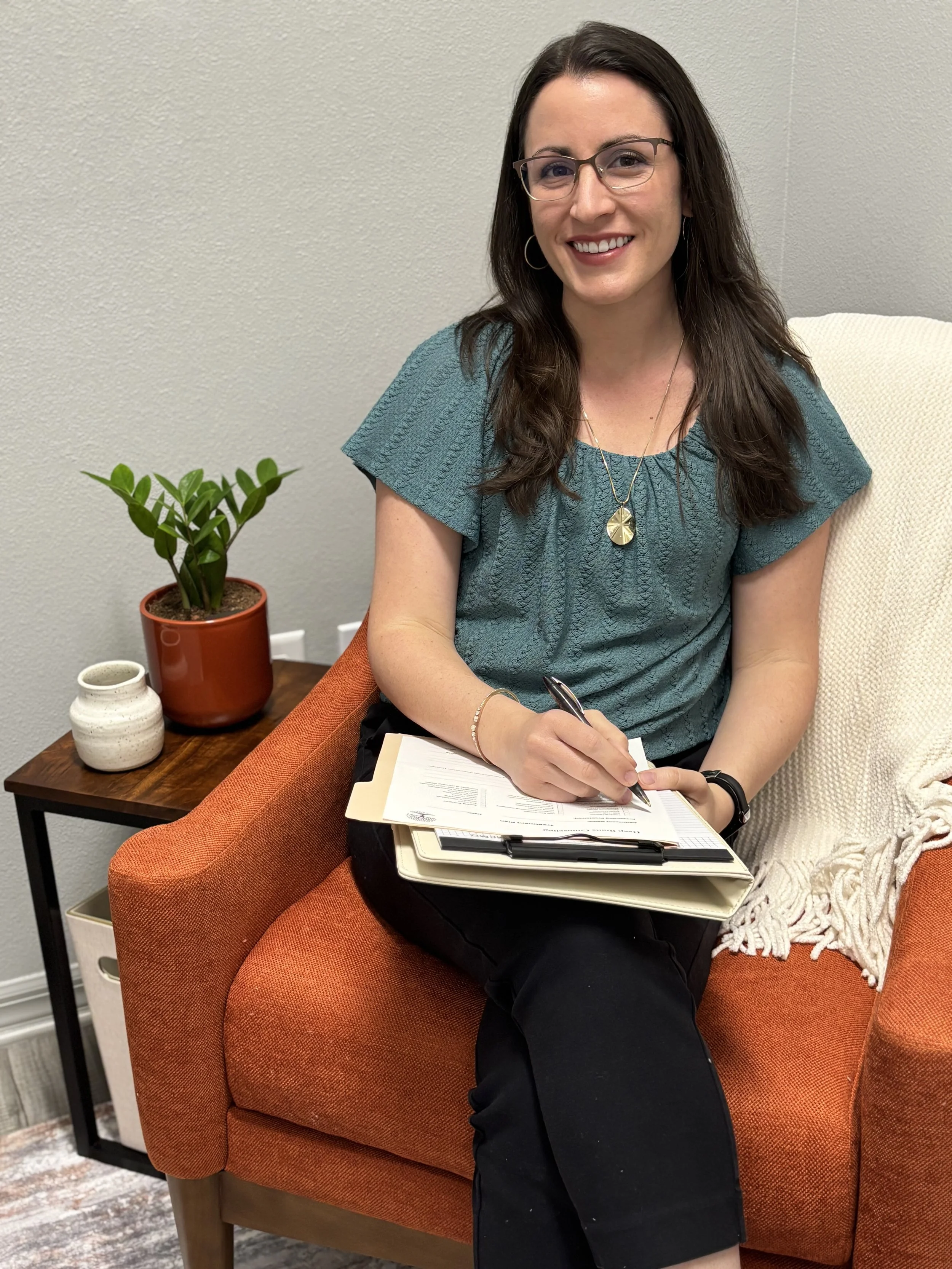A woman with glasses sitting on an orange armchair, smiling, holding a pen, and writing on documents on her lap in an office setting with a small table beside her holding a plant and a small vase.