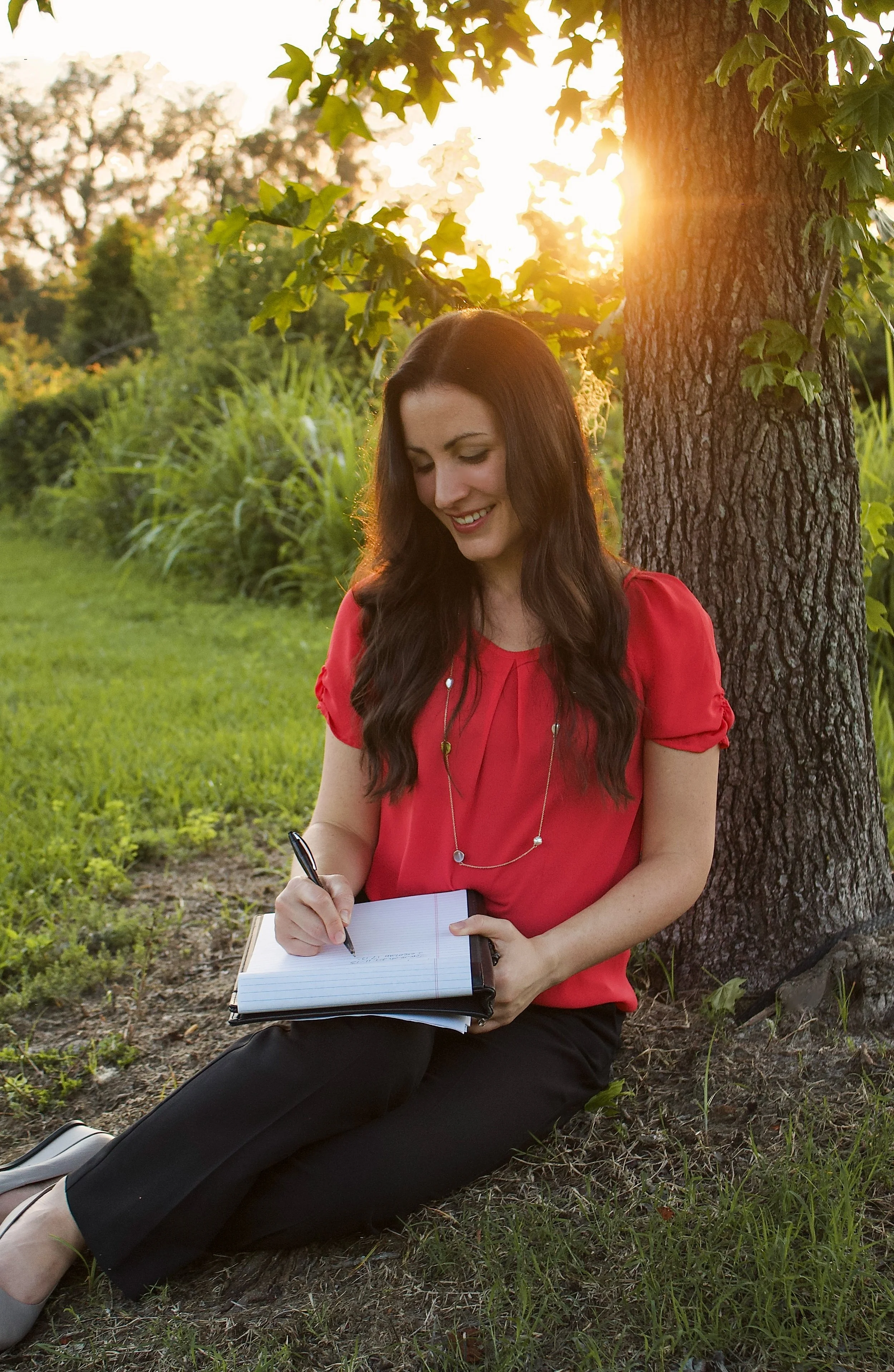 Woman sitting on the grass near a tree, writing in a notebook, with sunlight shining through the leaves at sunset.