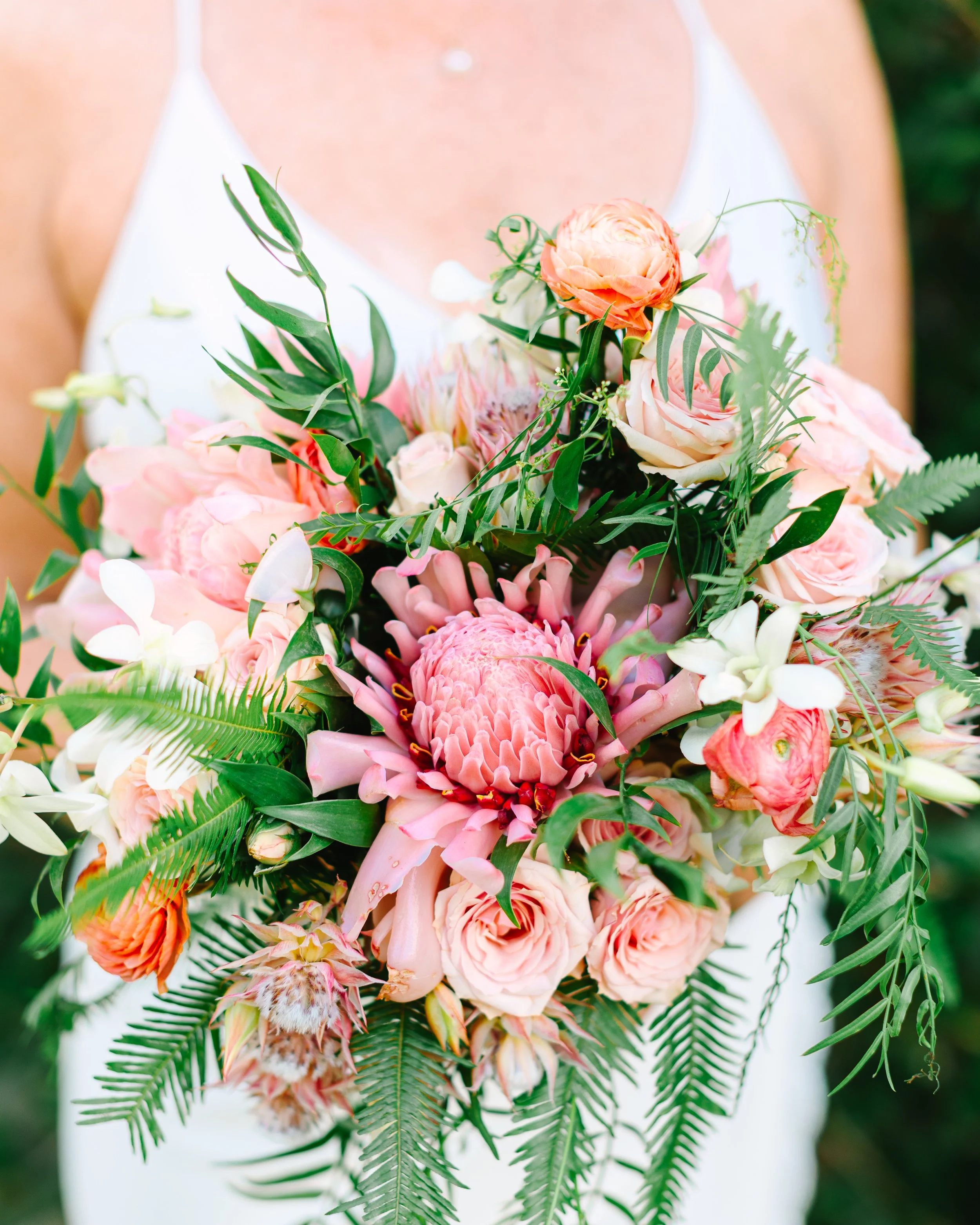 Close-up of a person holding a large bouquet of pink and white flowers with green leaves, dressed in a white sleeveless top.