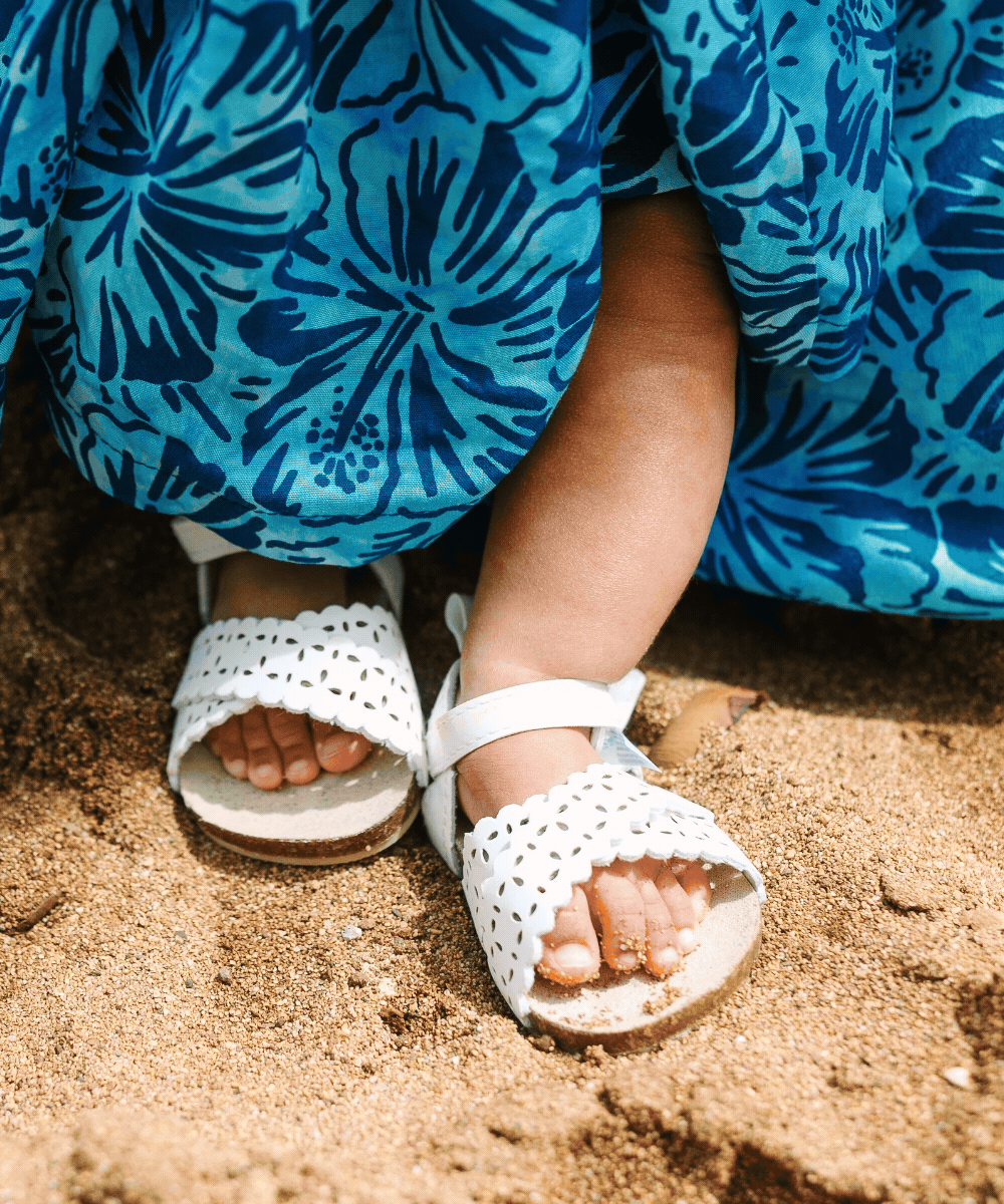 Child in a blue Hawaiian style dress and white sandals standing on sandy ground, with toes peeking out from sandals.