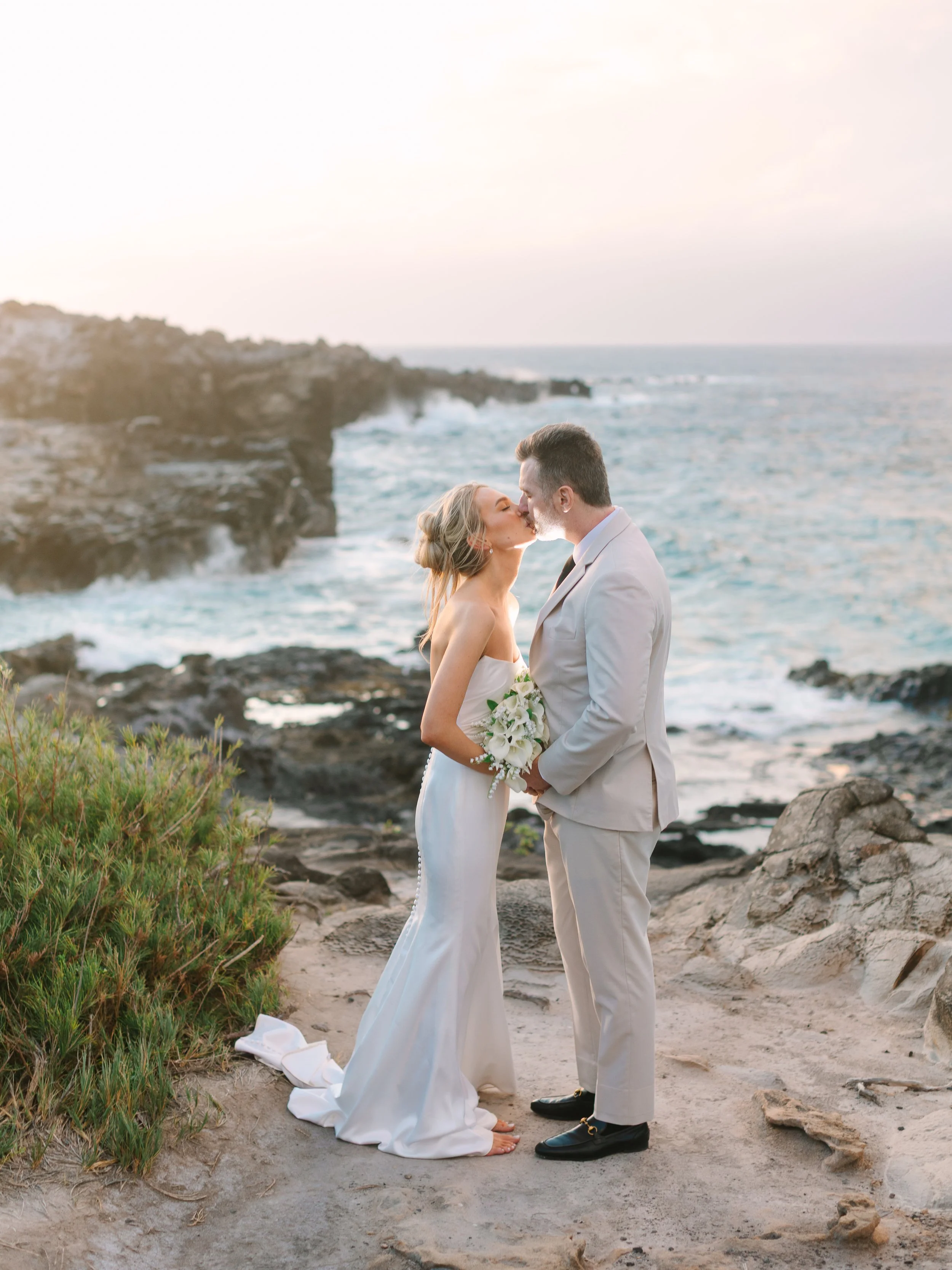 A bride and groom share a kiss on the beach at sunset, with ocean waves and rocky cliffs in the background.