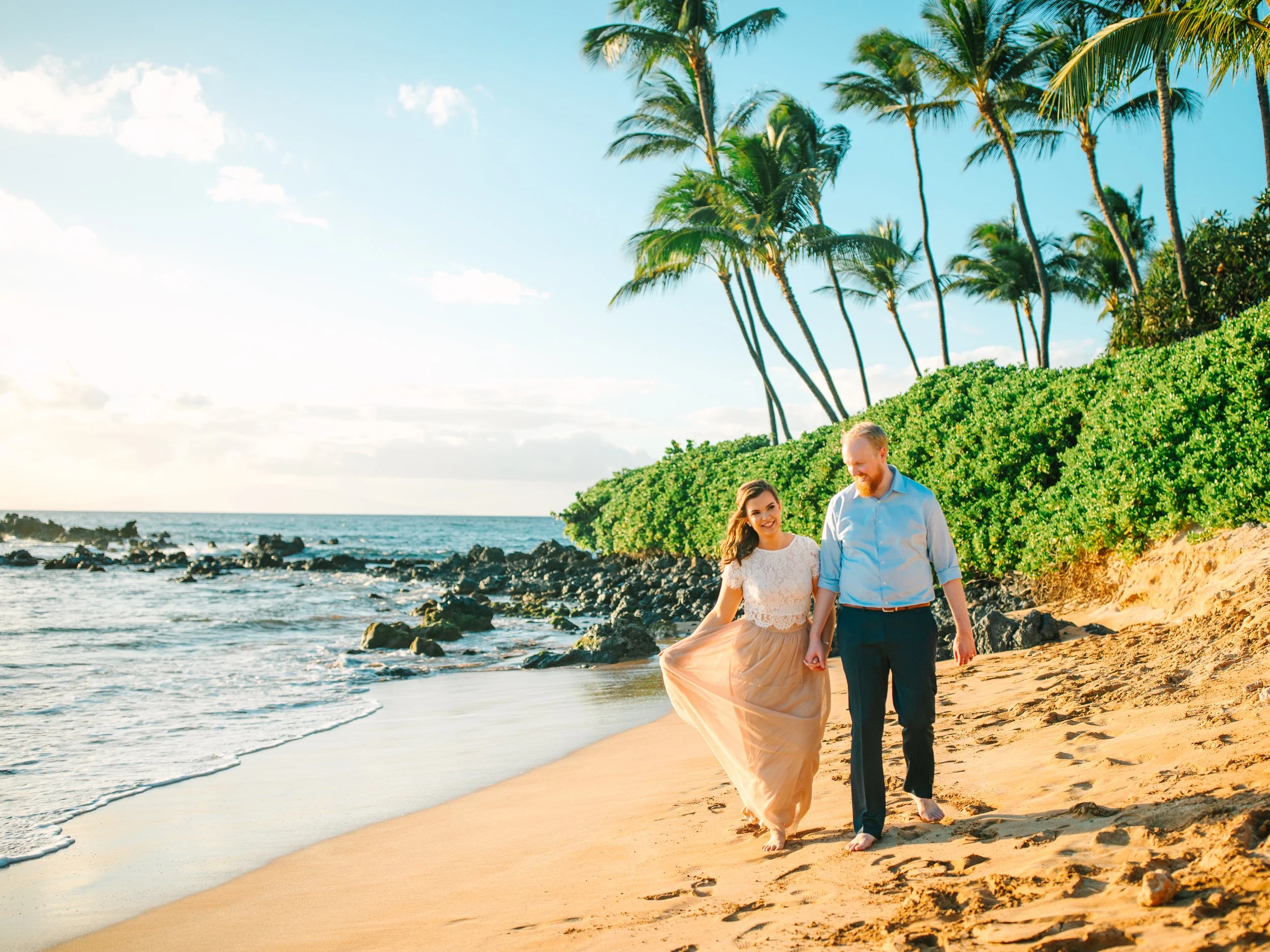 A couple walking hand in hand on a sandy beach, with palm trees and rocks along the shoreline, under a partly cloudy sky.