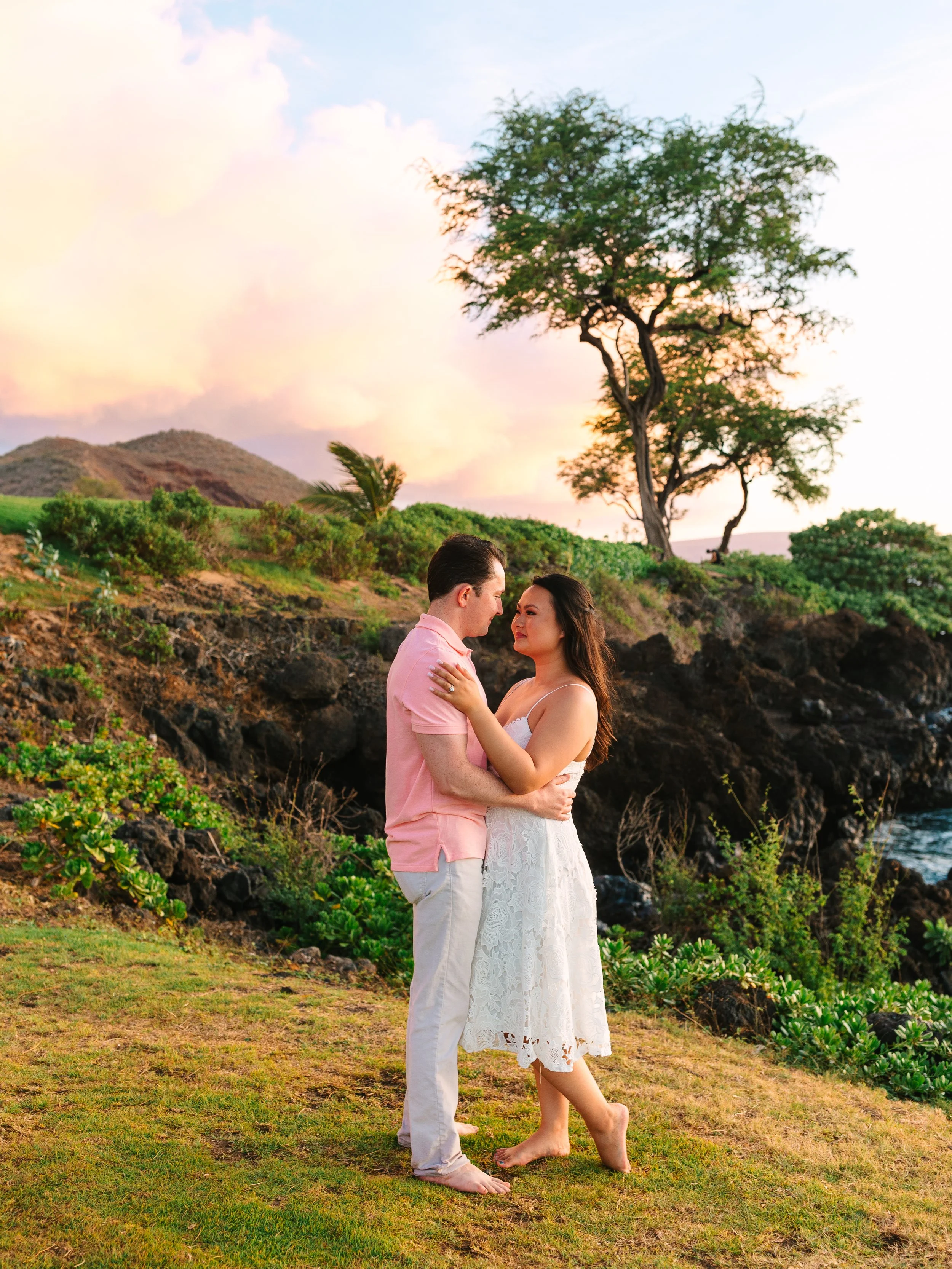 A couple standing close together, embracing on a grassy area near the coast during sunset, surrounded by lush greenery and a large tree, with mountains and a colorful sky in the background.