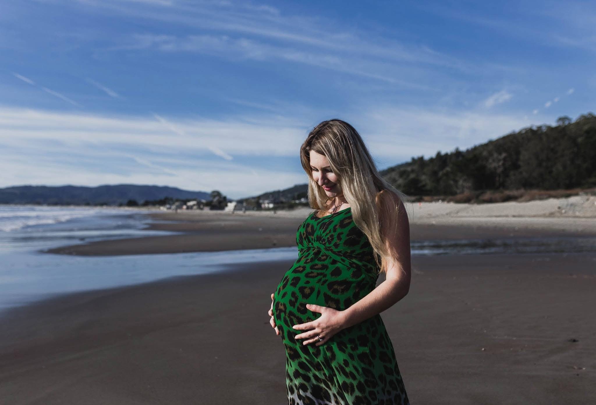 Pregnant woman in a green and black leopard print dress standing on a beach, holding her belly and looking down.