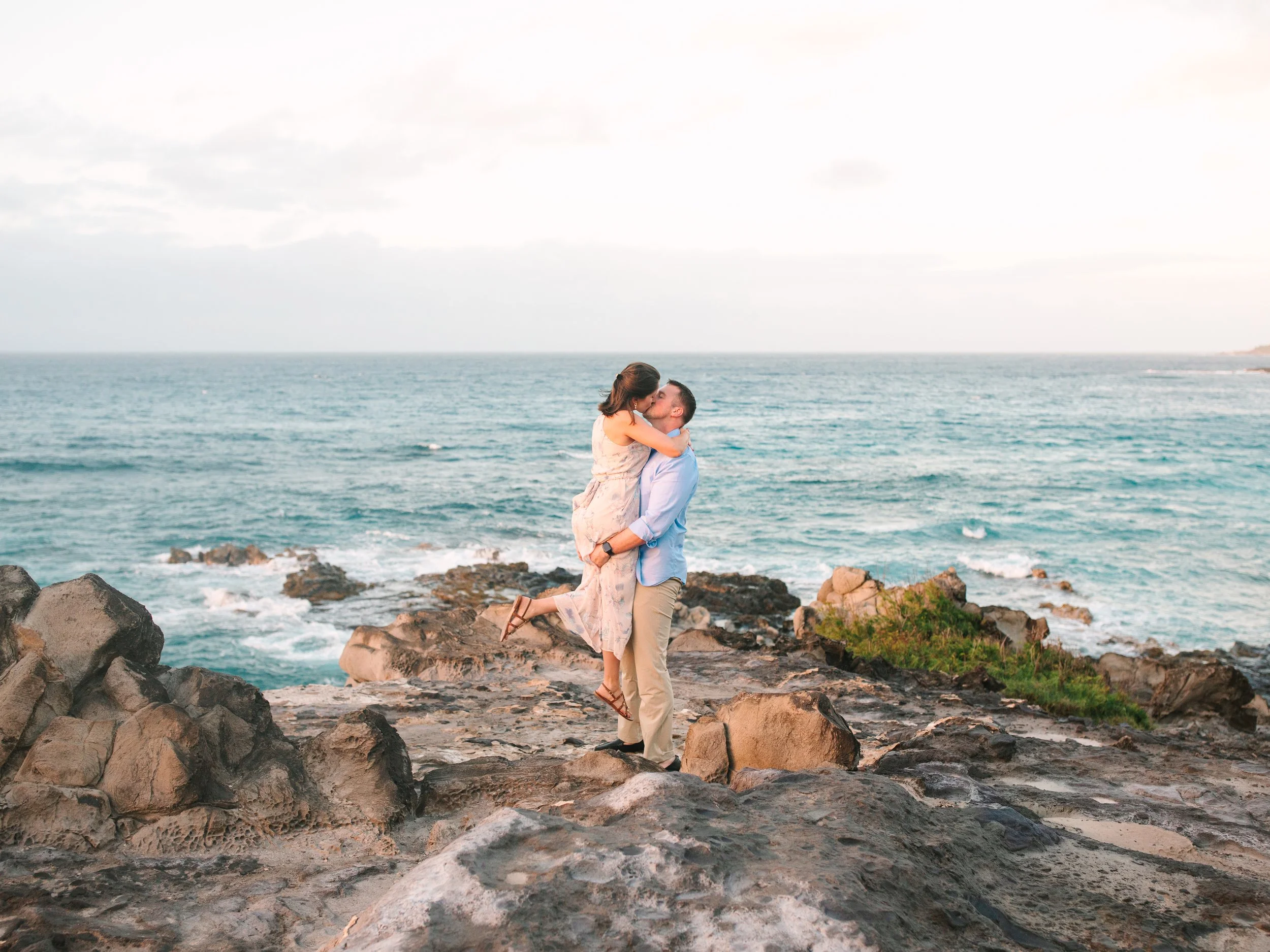 A couple kissing on a rocky beach, with the ocean and a partly cloudy sky in the background.