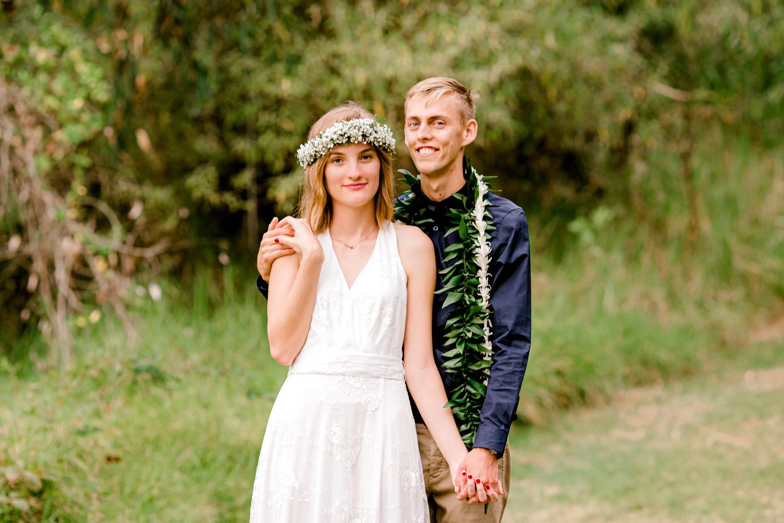 A young couple dressed in wedding attire standing outdoors in a green natural setting, holding hands, with the man wearing a flower lei and the woman wearing a floral headband.