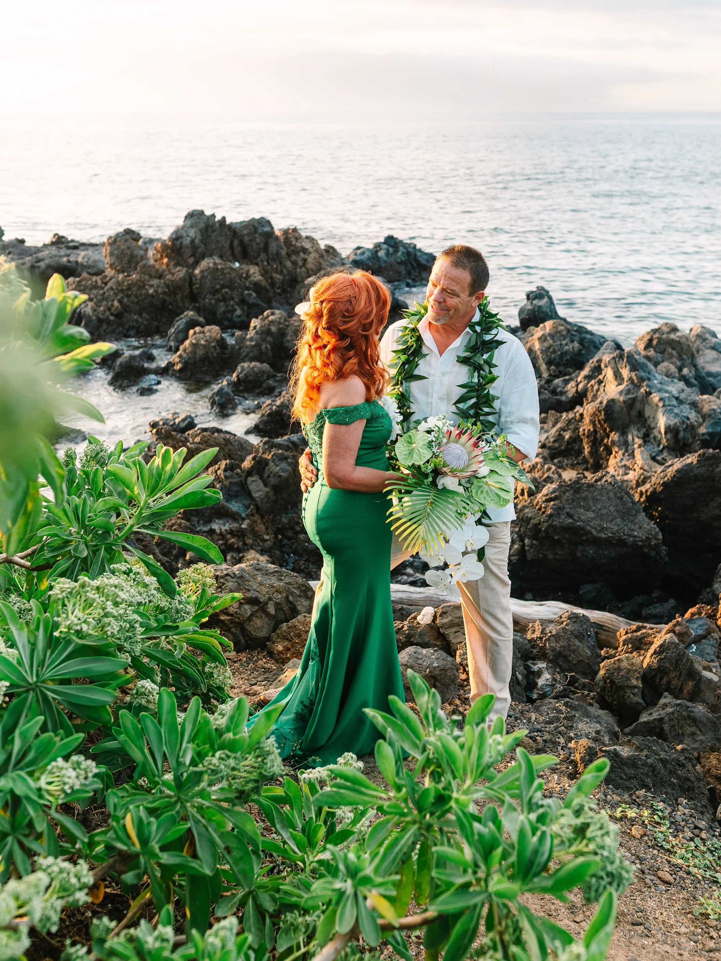 A couple exchanging vows on a rocky beach with the ocean in the background, surrounded by greenery, during sunset.