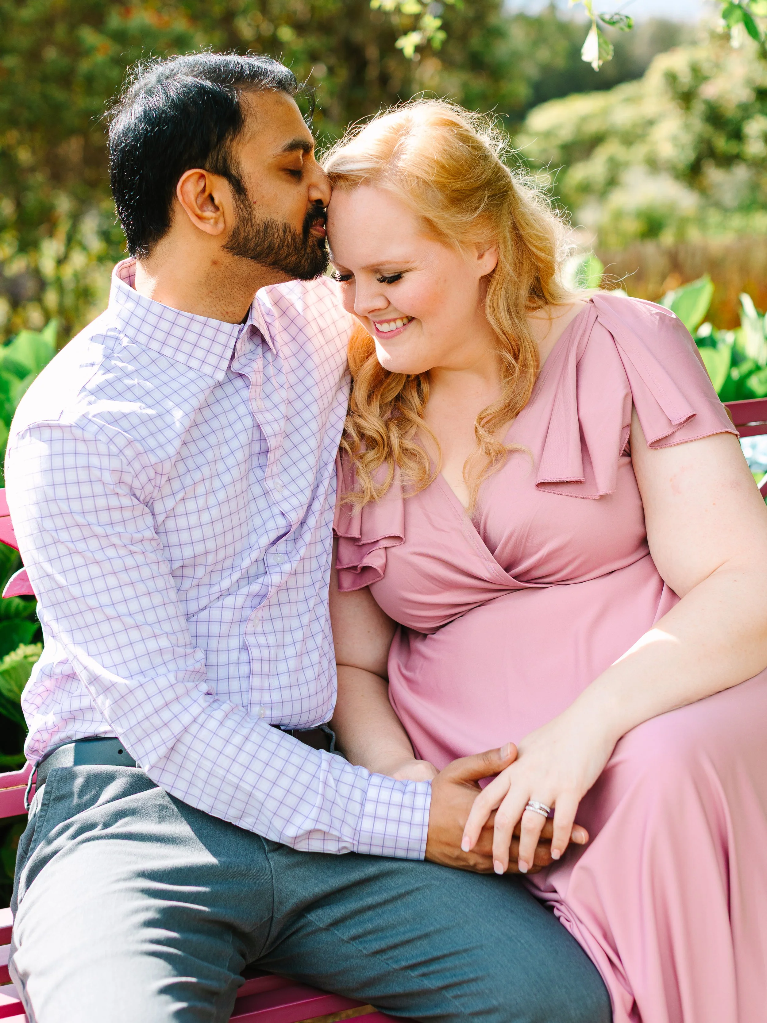 A couple sitting on a park bench, with the man kissing the woman's forehead, both smiling.