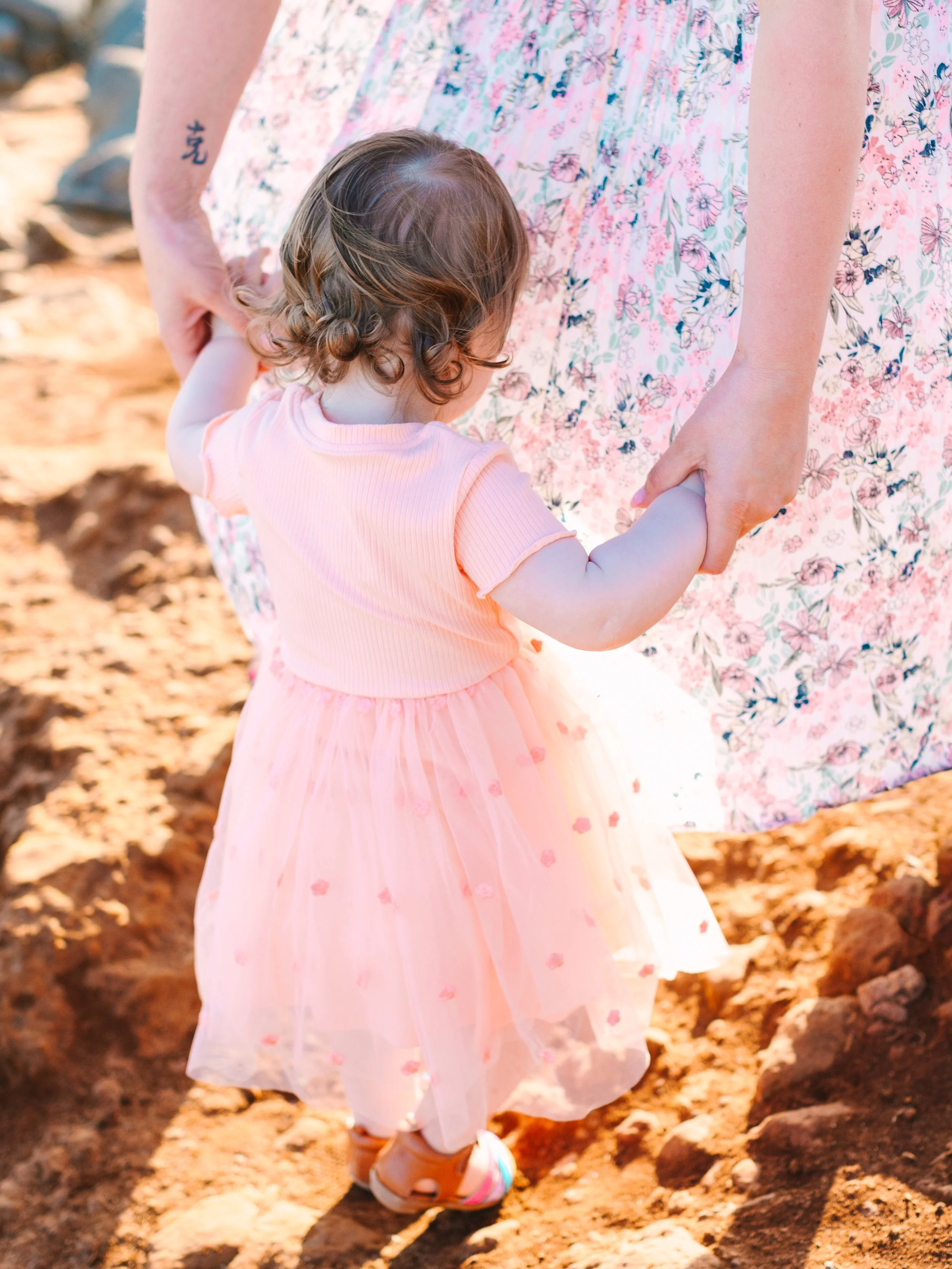 A young girl in a pink dress holding hands with an adult woman in a floral dress outdoors on a dirt path.