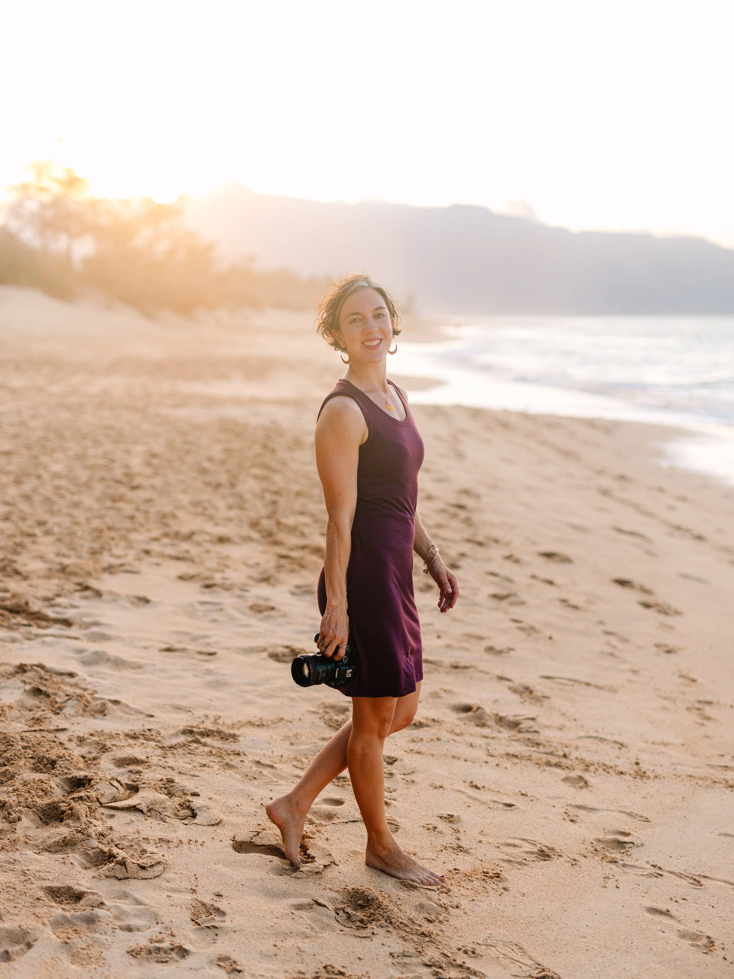 A woman in a sleeveless purple dress walking barefoot on a sandy beach at sunset, holding a camera in her right hand and smiling at the camera.