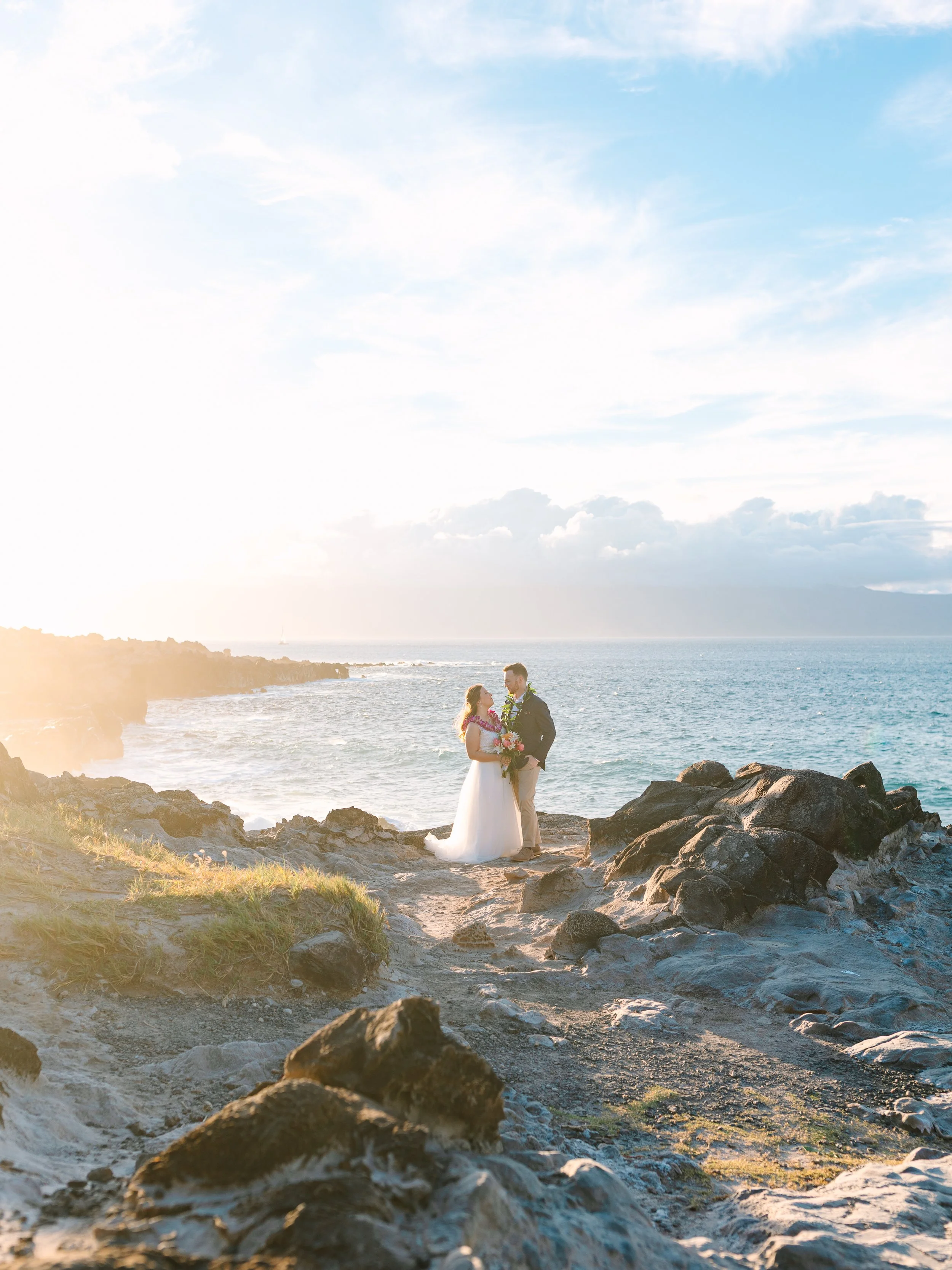 A couple in wedding attire standing on a rocky coastal area, exchanging vows or sharing a moment at sunset with the ocean and sky in the background.