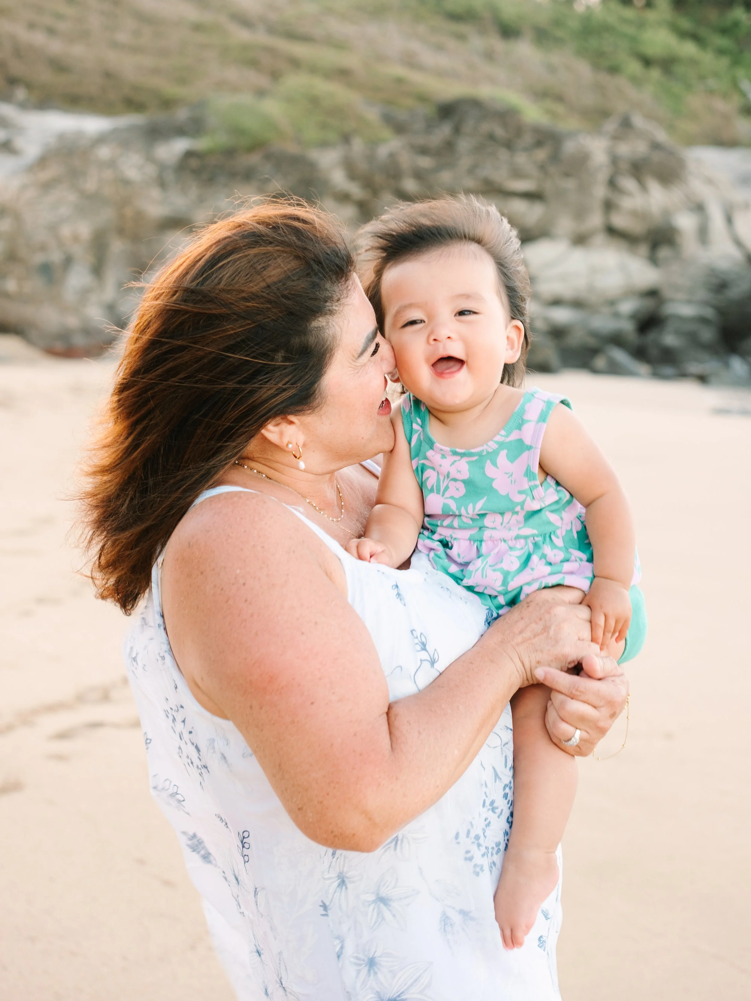 A woman holding a smiling baby girl on a beach with rocks and sand in the background.