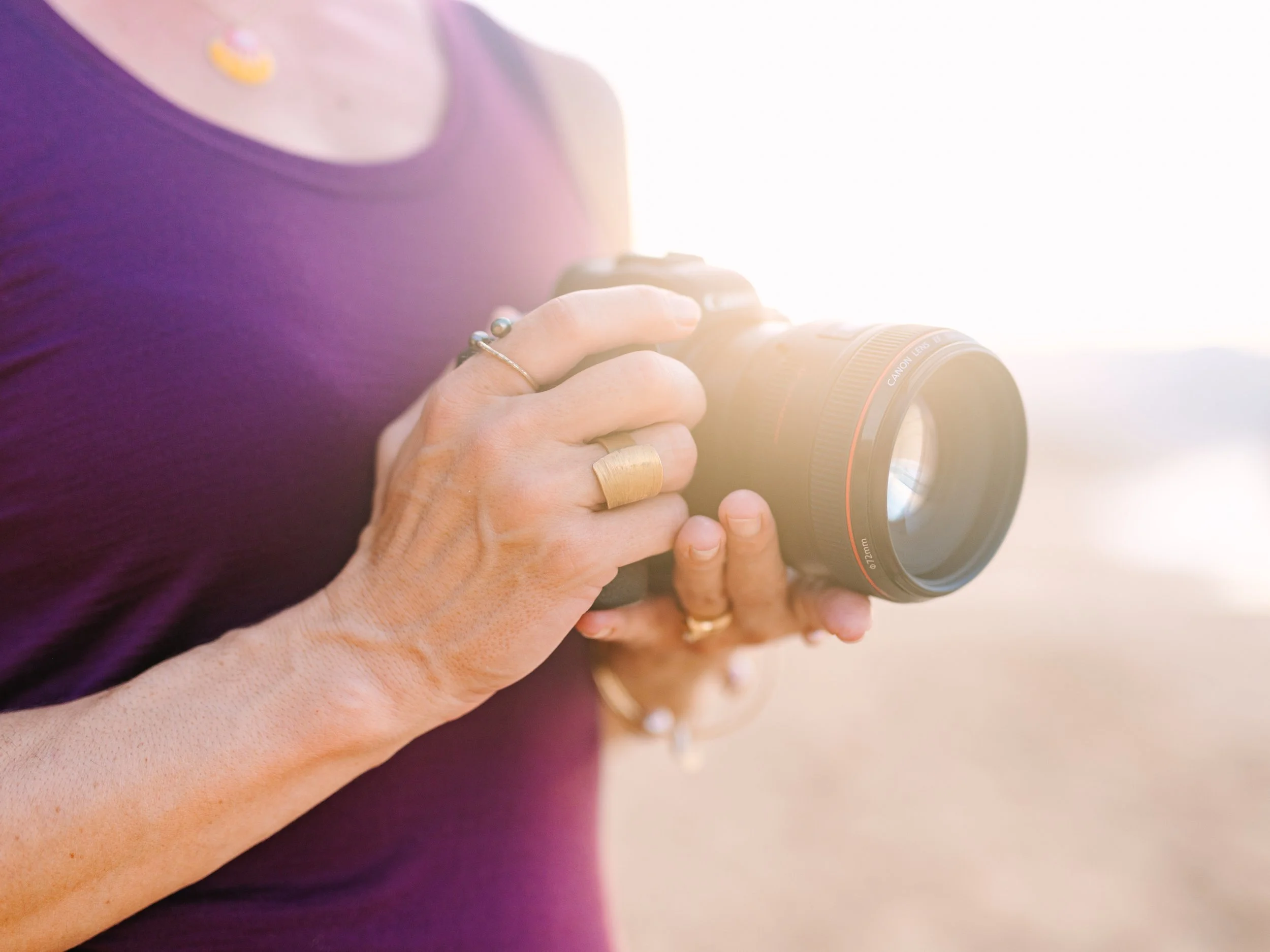 Person holding a professional camera with both hands, wearing a purple top and jewelry, with sunlight in the background.
