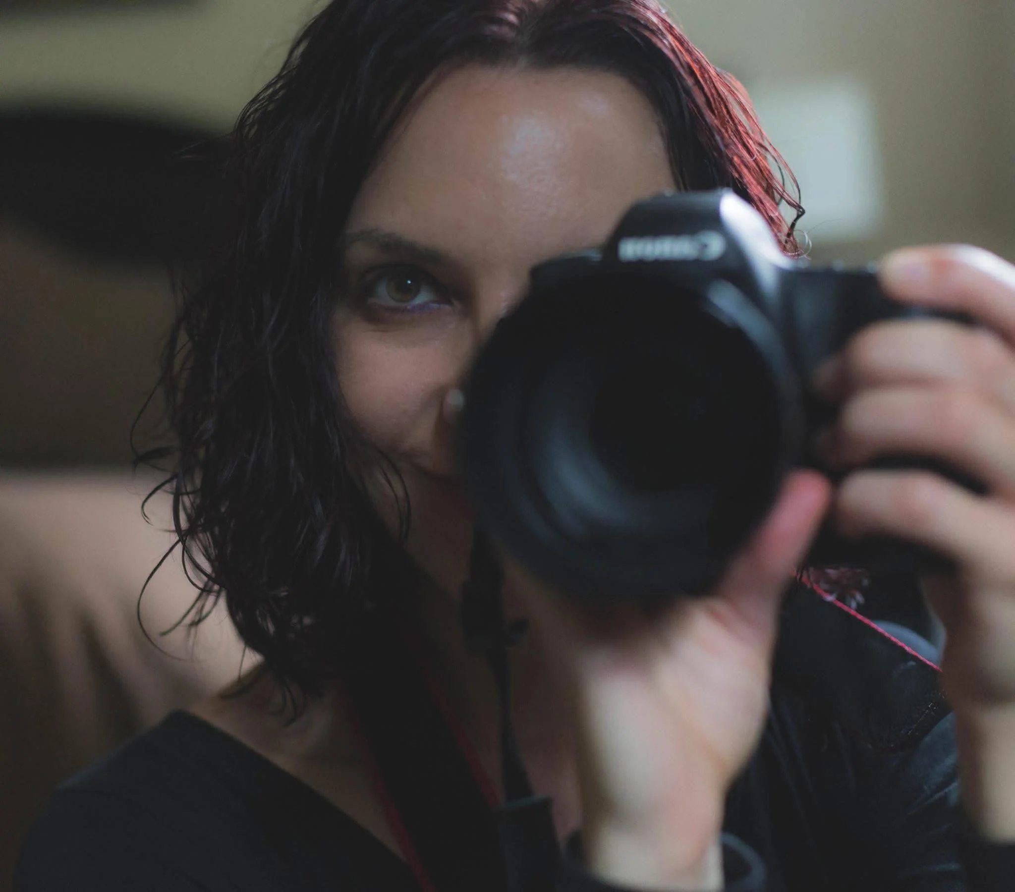 A woman with dark wavy hair holding a Canon camera, taking a selfie in front of a mirror.