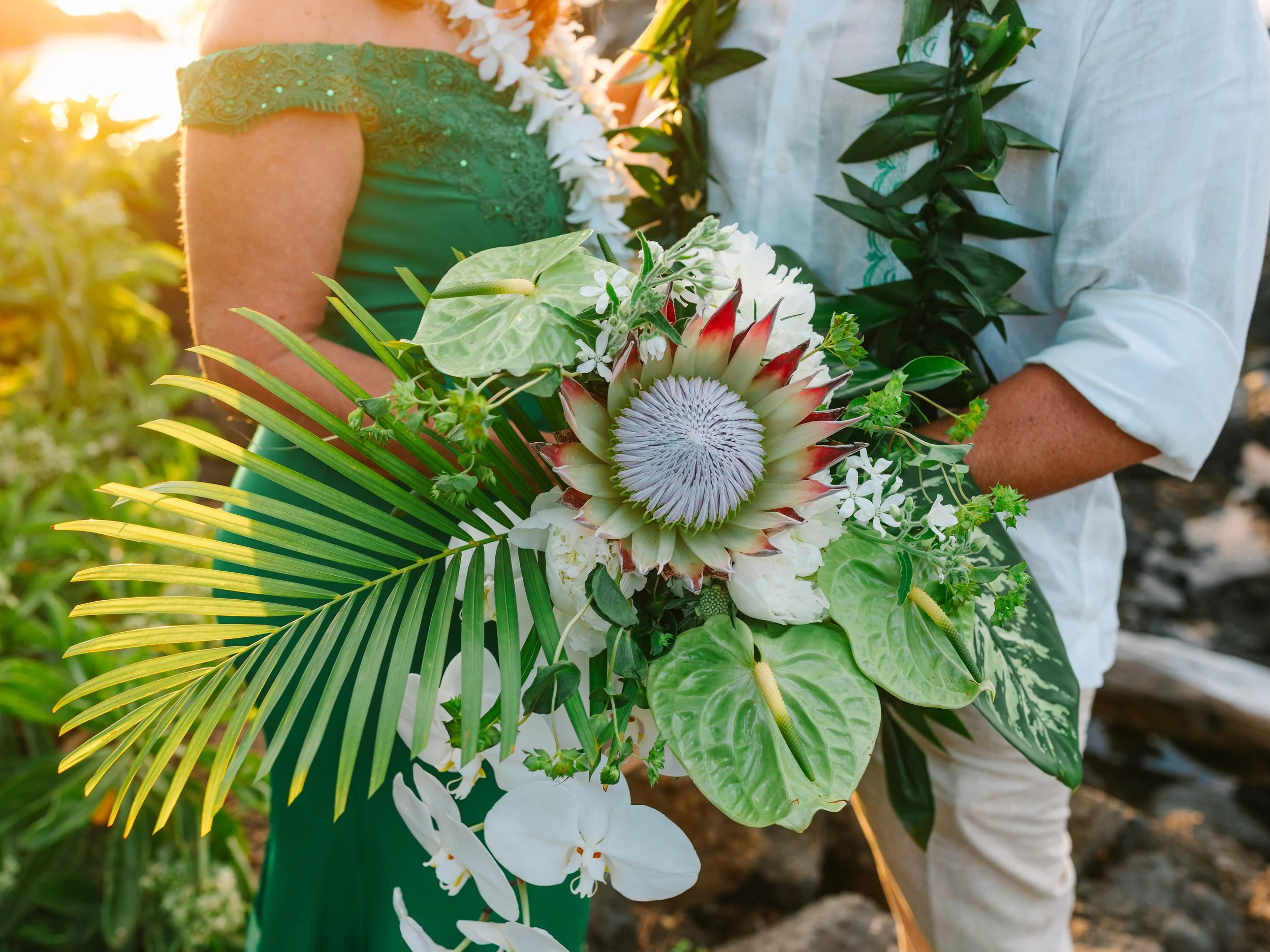 Couple holding a tropical bouquet of flowers including a large protea, white orchids, anthuriums, and various green leaves during sunset.