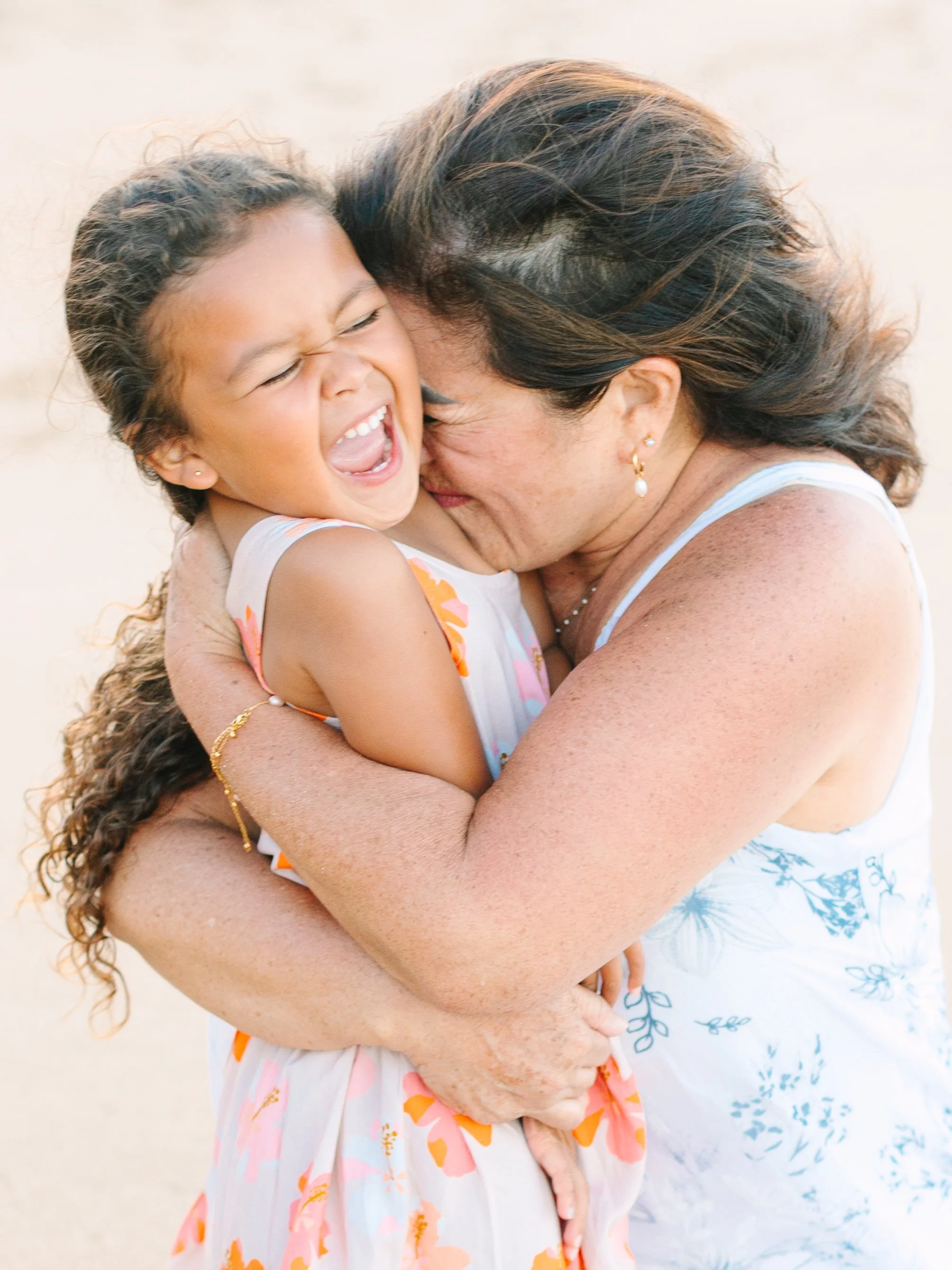 A woman and a young girl share a joyful hug on the beach, both smiling and laughing happily.