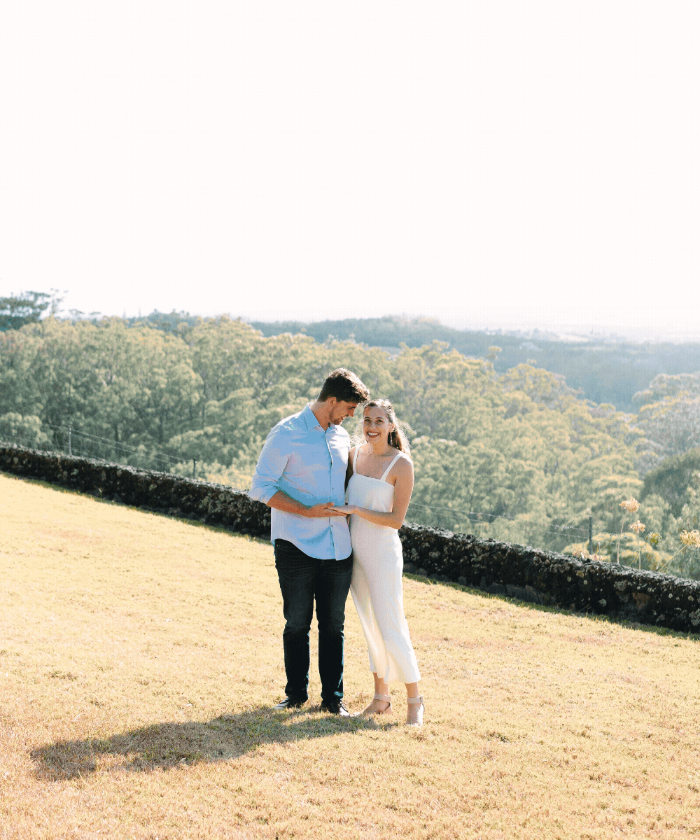 A young couple stands outdoors on a sunny day, holding hands and smiling. The man wears a light blue shirt and dark pants, while the woman wears a white summer dress. They are standing on a grassy area with a scenic background of trees and hills.