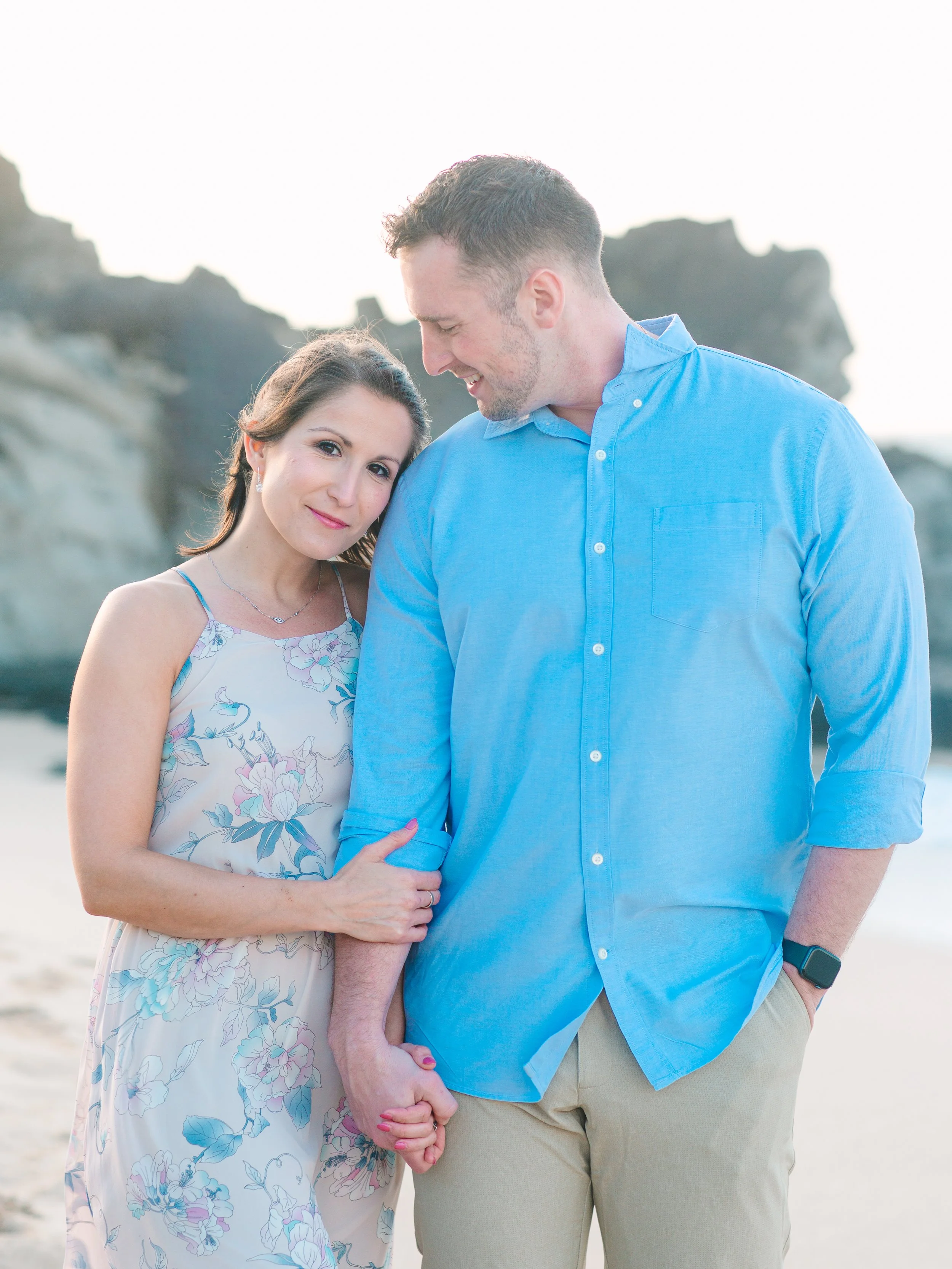 A couple holding hands and standing on a beach, with rocky cliffs in the background, during sunset.