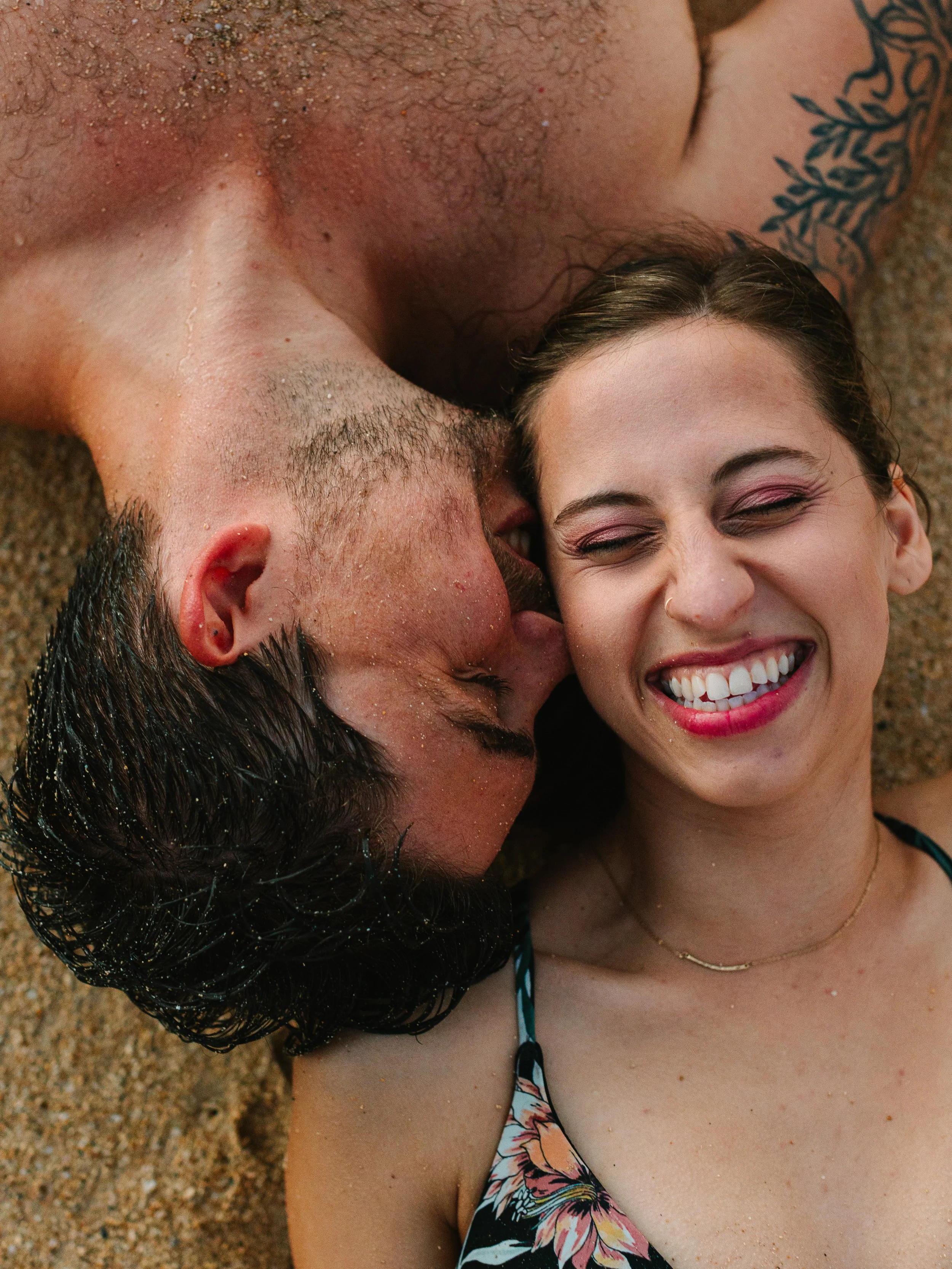 A young couple lying on the sandy beach, smiling and close to each other, with the man kissing the woman's cheek.