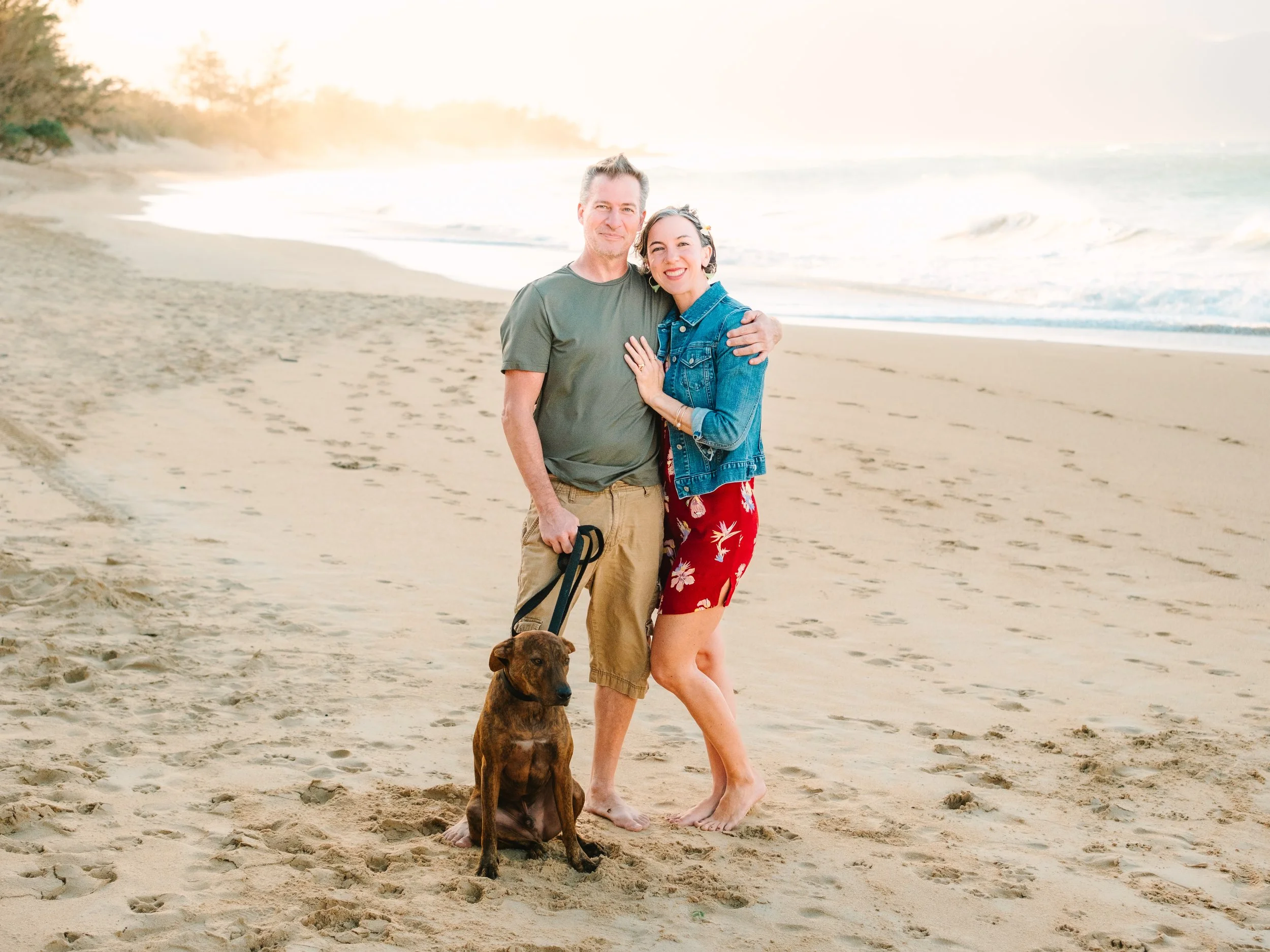A couple with a dog standing on a sandy beach at sunset, smiling and embracing each other.
