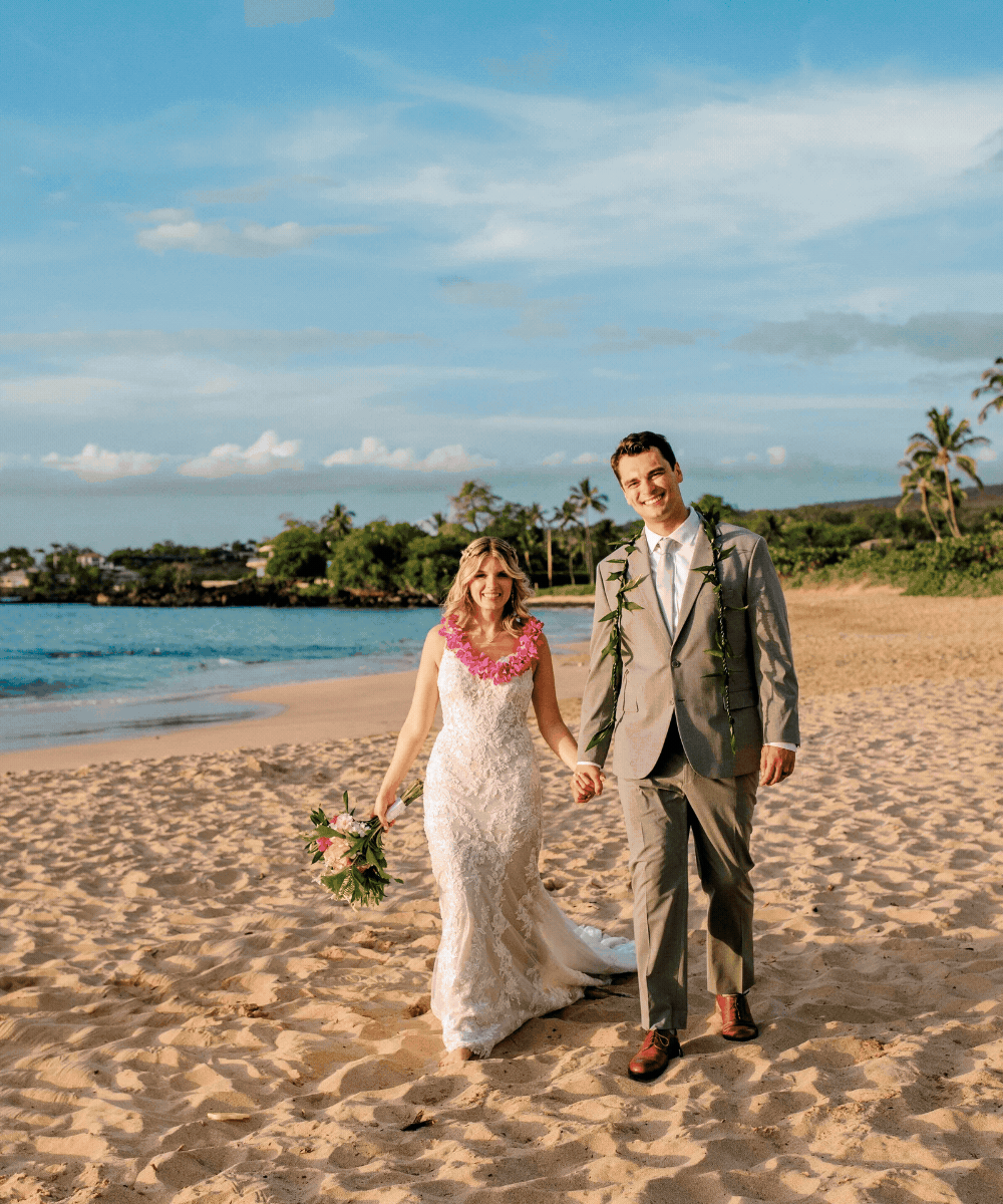 A bride and groom walking hand in hand on a sandy beach during sunset, with palm trees and a calm ocean in the background.