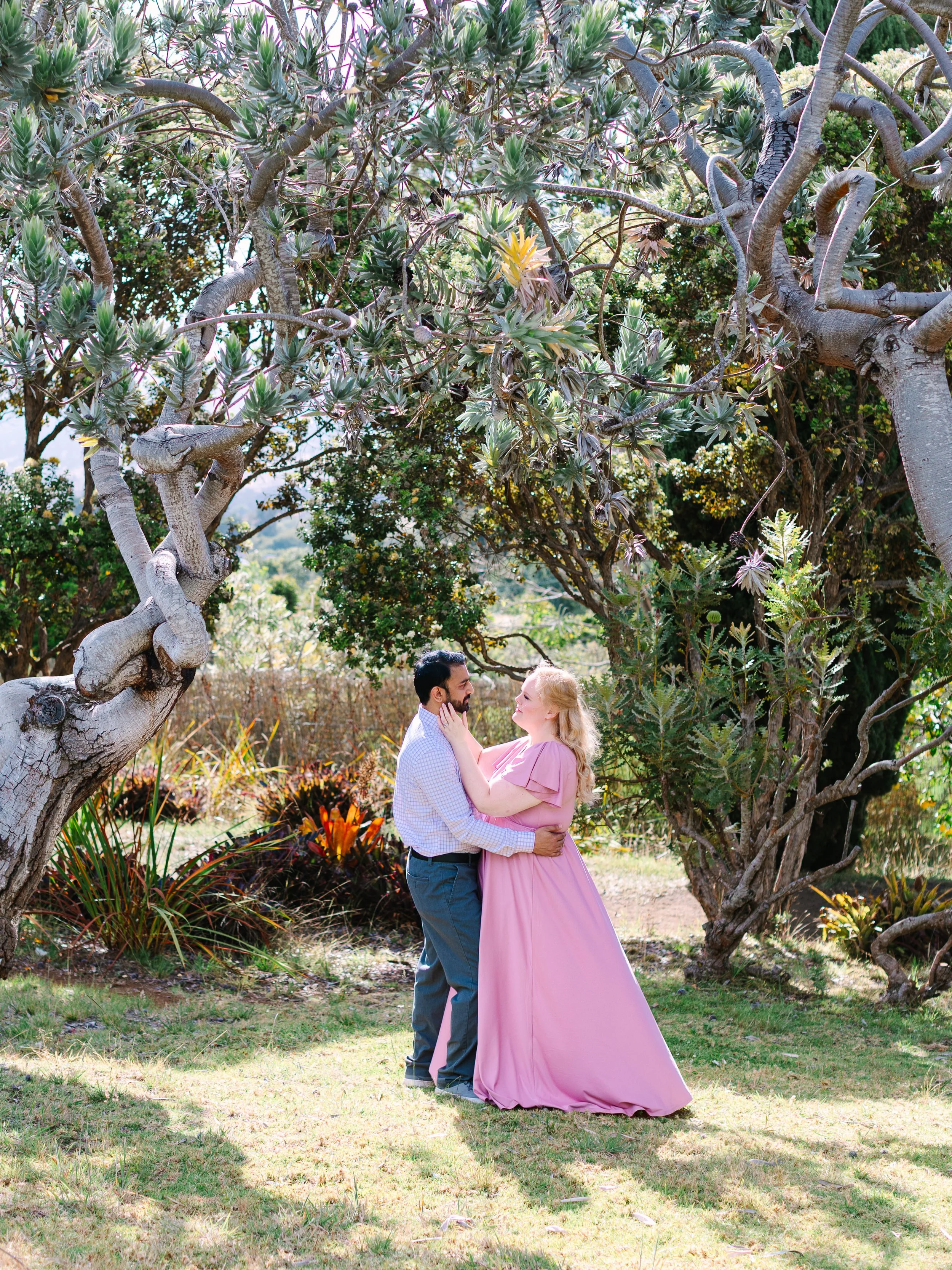 A couple embraces in a lush outdoor setting with large trees and vibrant plants, woman in long pink dress, man in light-colored shirt and pants.