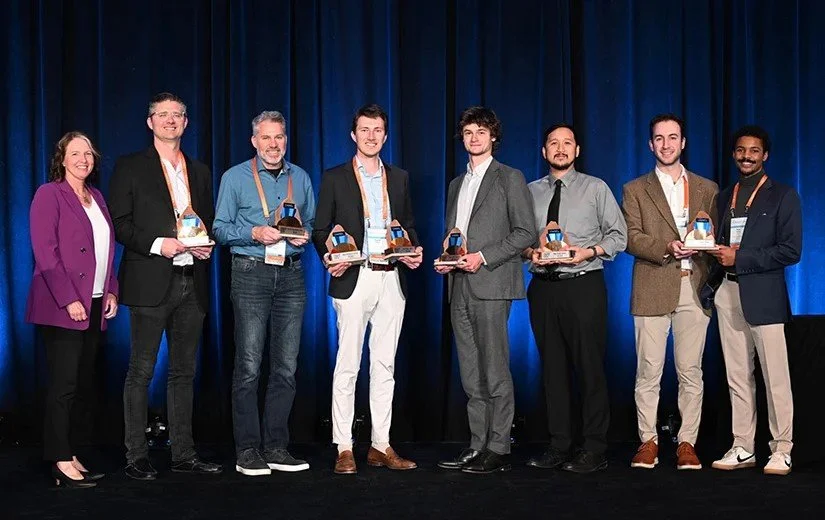 Group of eight people standing on stage holding awards, with a blue curtain background, at an awards ceremony or conference.