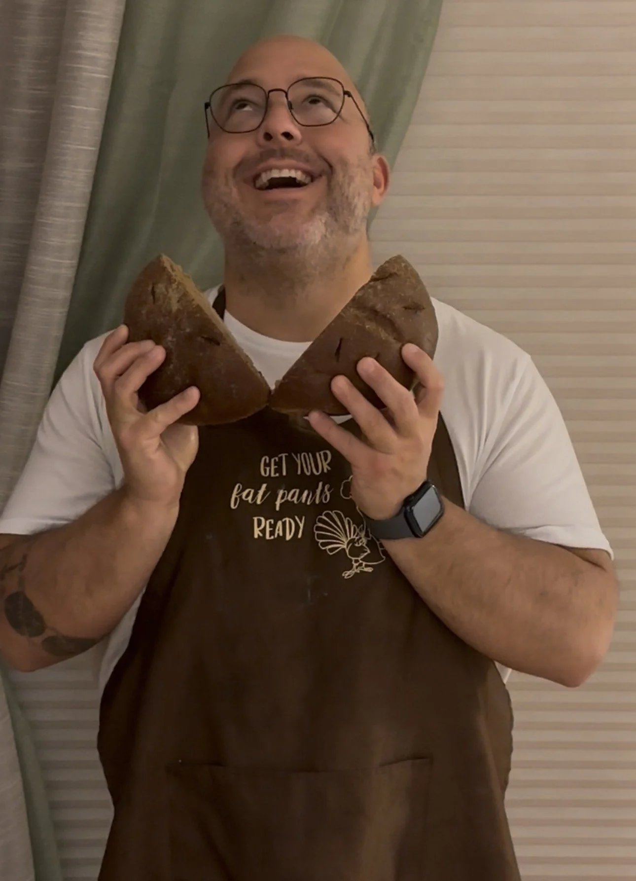 A man is smiling and looking up while holding two halves of a large loaf of bread. He is wearing glasses, a white T-shirt, a dark brown apron with the phrase "Get your fat pants ready" and a graphic, a black wristwatch, and has tattoos on his right arm. The background includes curtains and horizontal blinds.