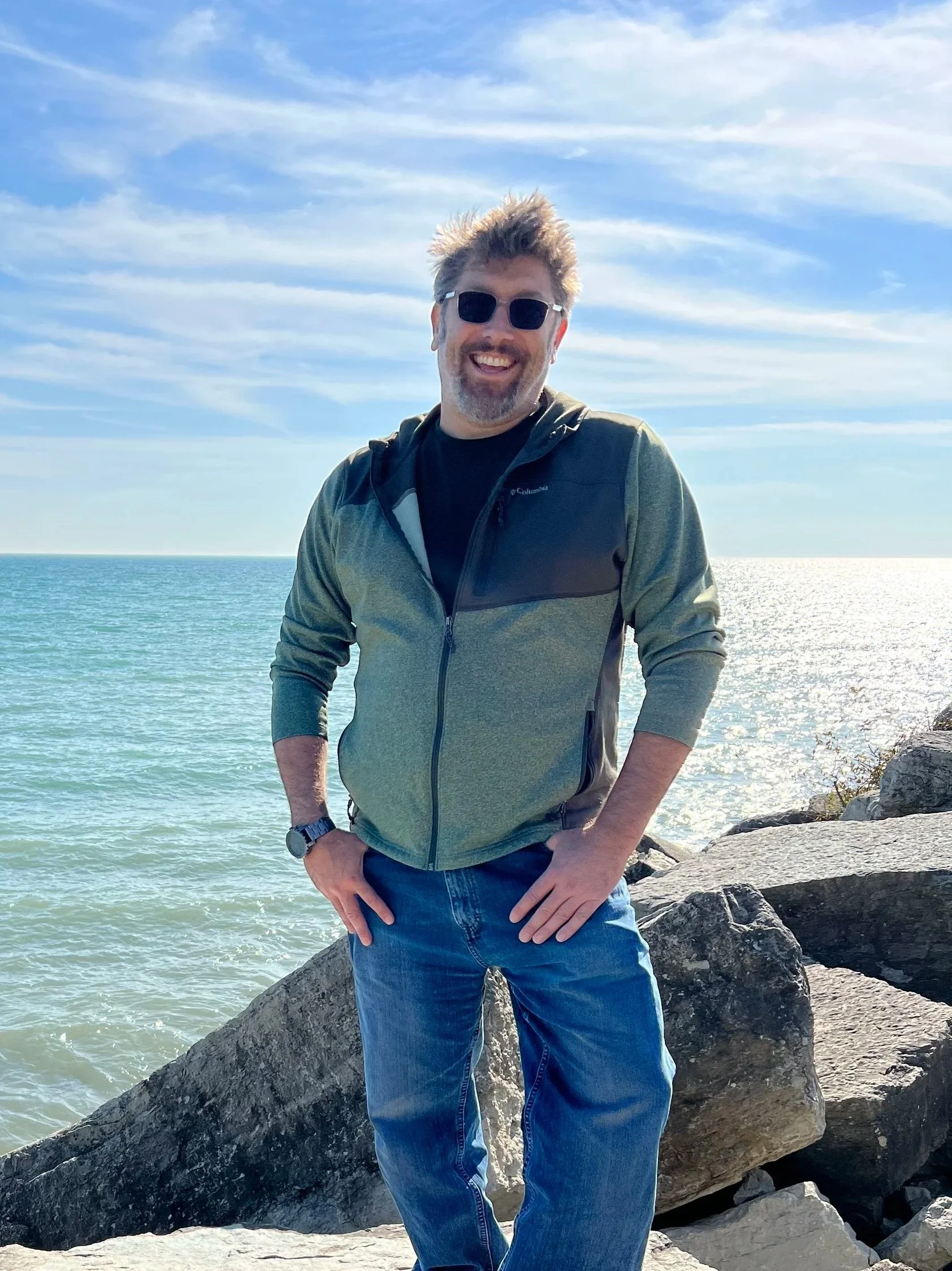 A man stands on rocks by the ocean, wearing sunglasses, a green and black jacket, and jeans, smiling at the camera with the sea and a partly cloudy sky in the background.