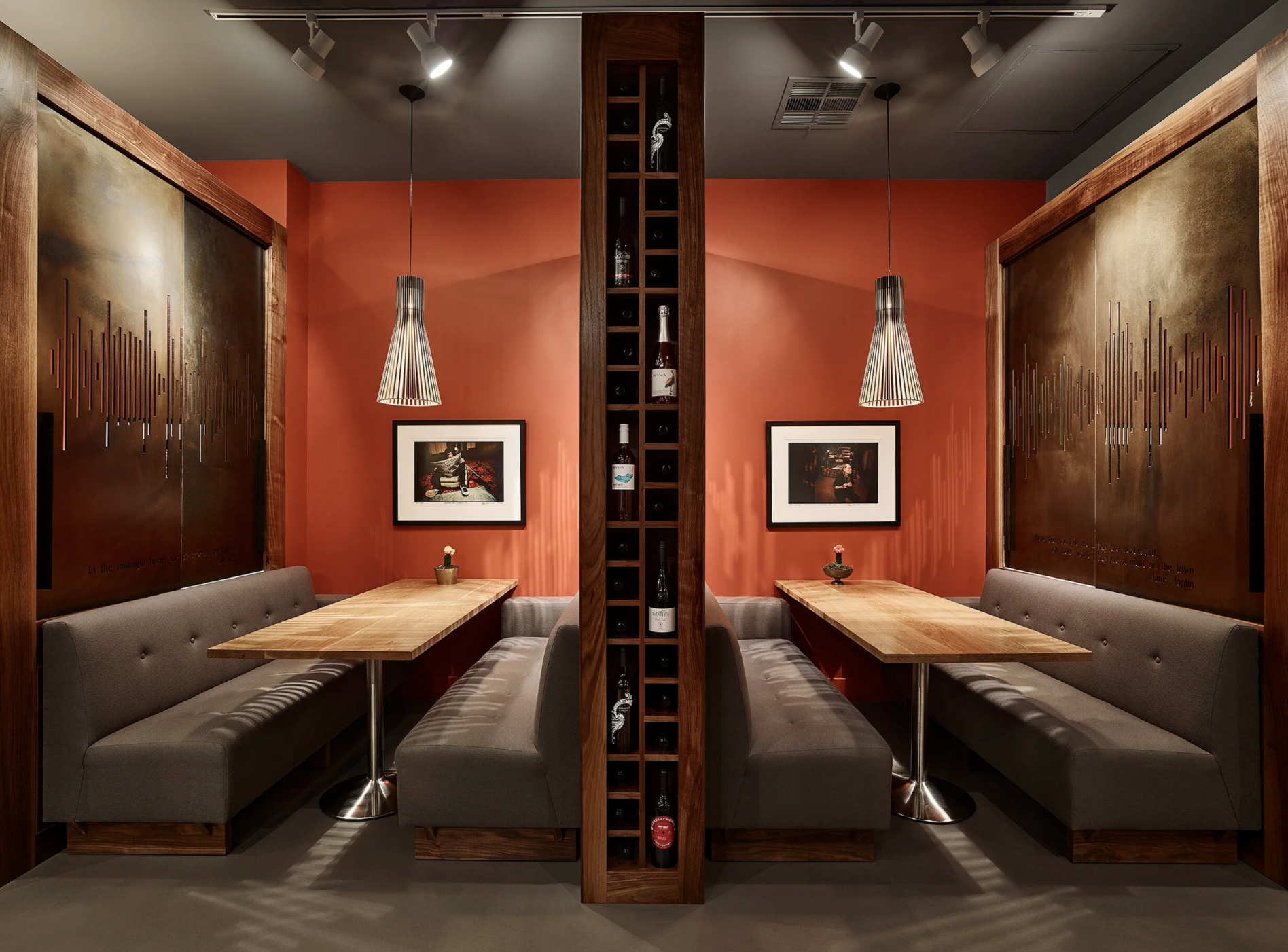 Symmetrical dining booth for four, separated by a wooden wine rack, with orange walls, framed photographs, and pendant lights.