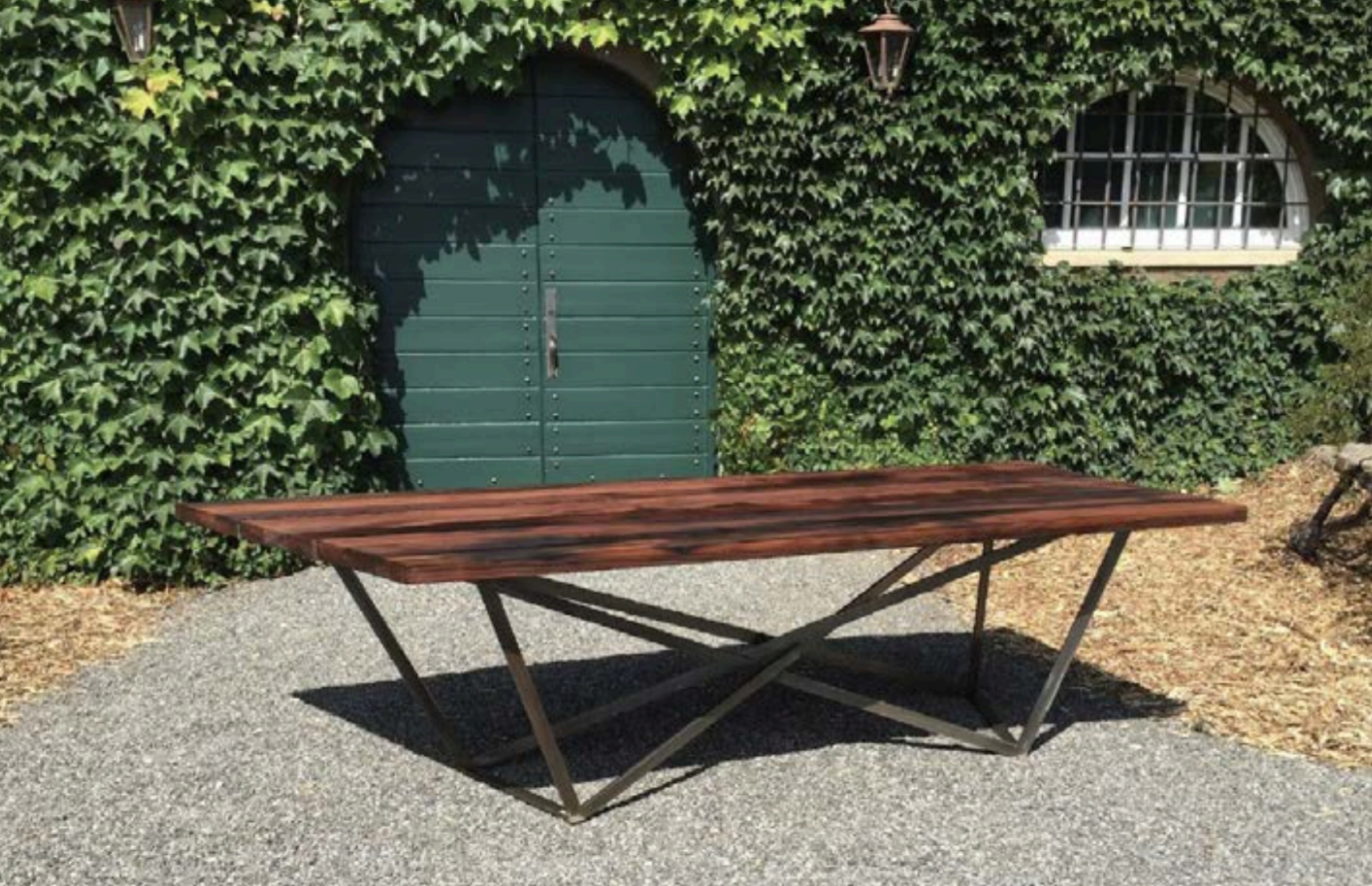 A wooden outdoor table with a metal base on a gravel surface, set against a background of a green ivy-covered wall and a green arched door.