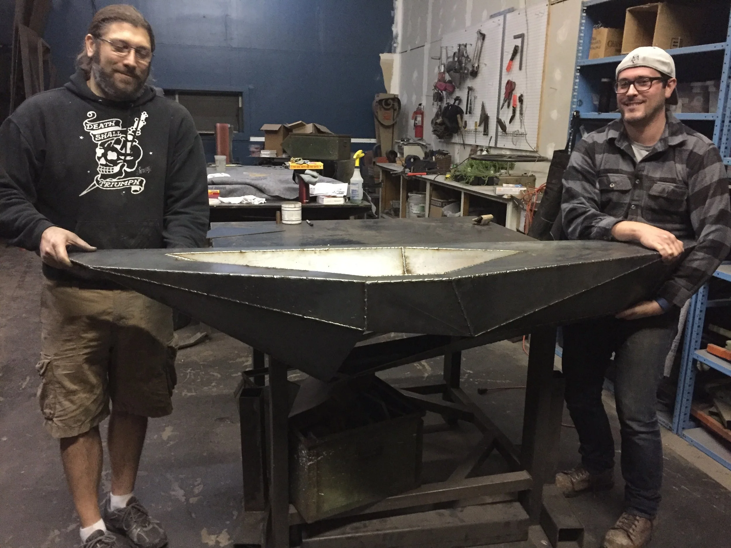 Two men holding a custom welded sink in a workshop with tools and shelves in the background.