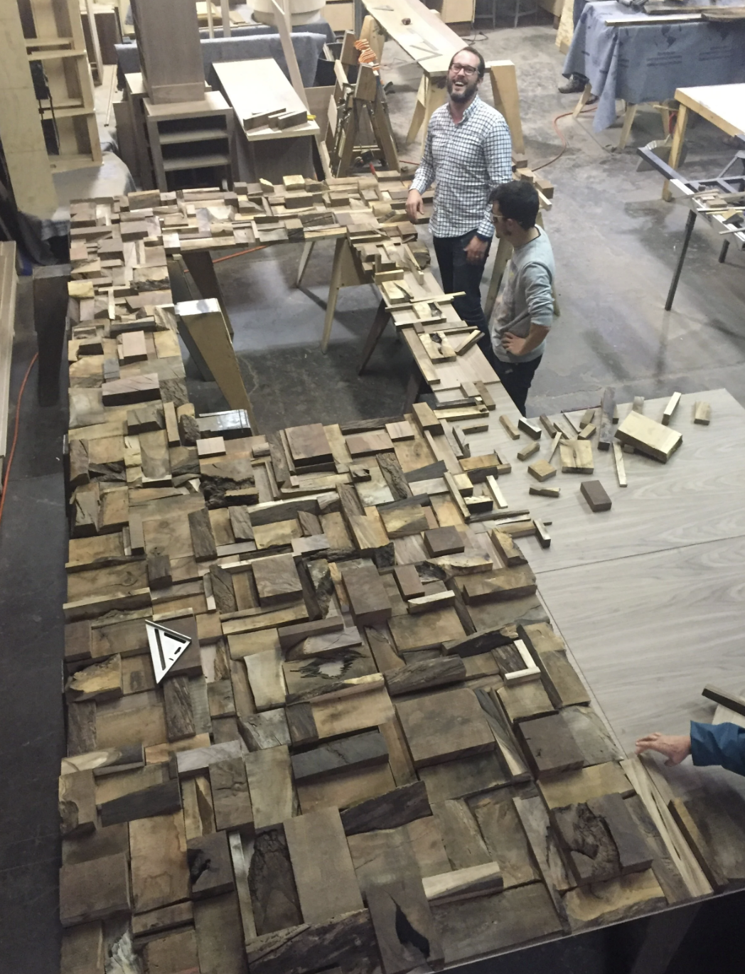 Two men standing at a woodworking workshop surrounded by various pieces of wood, with a table displaying a large, intricate wood mosaic or collage made from different wood pieces.