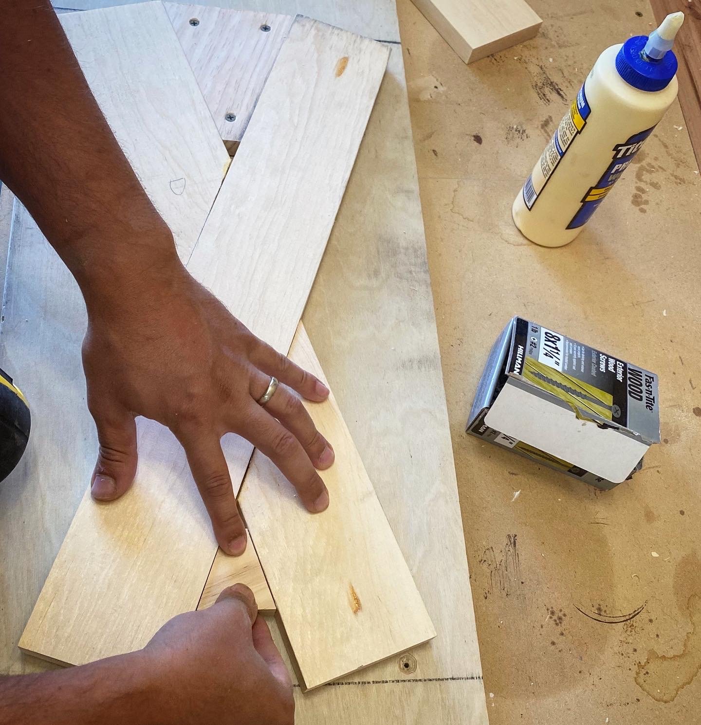 Person assembling wooden pieces on a workbench with glue and a box of saw blades nearby.