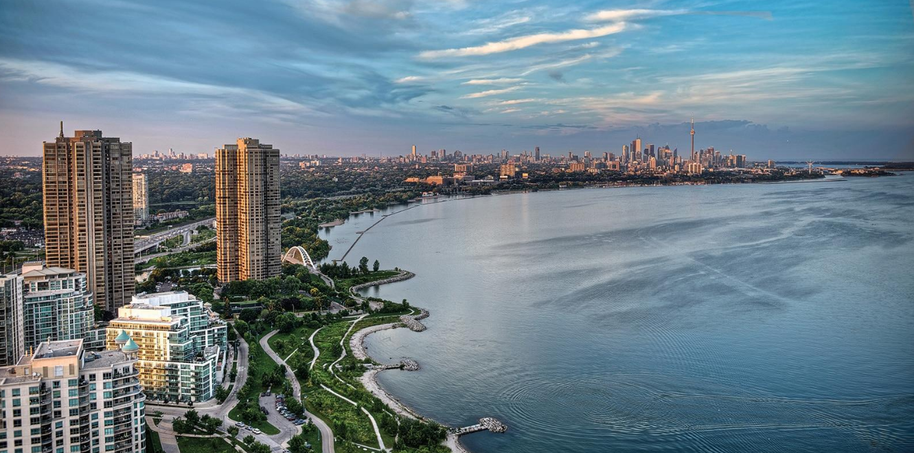 A city skyline with tall buildings along a large body of water, green parks in the foreground, and a partly cloudy sky.