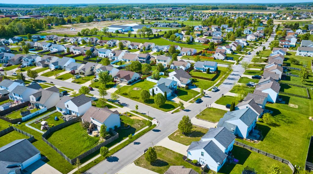 Aerial view of a suburban neighborhood with single-family homes, manicured lawns, trees, and streets in a grid pattern.