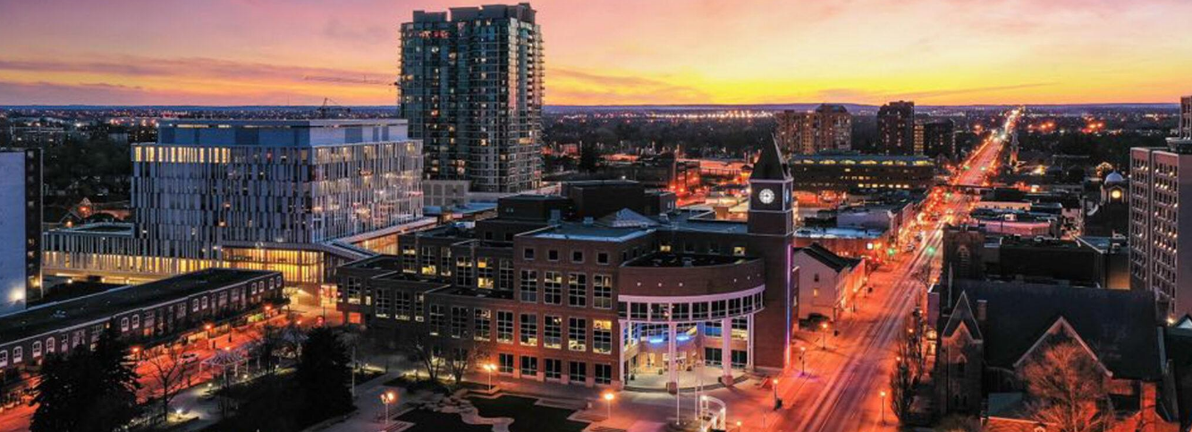 Cityscape at sunset with a mix of modern and historic buildings, illuminated streets, and a clock tower in the foreground.