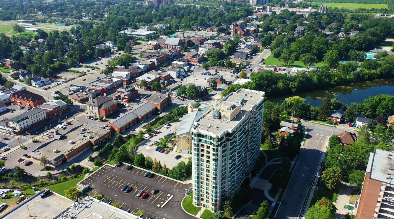 Aerial view of a city showing a large high-rise building, a parking lot, a river, and a mix of residential and commercial buildings surrounded by green trees.
