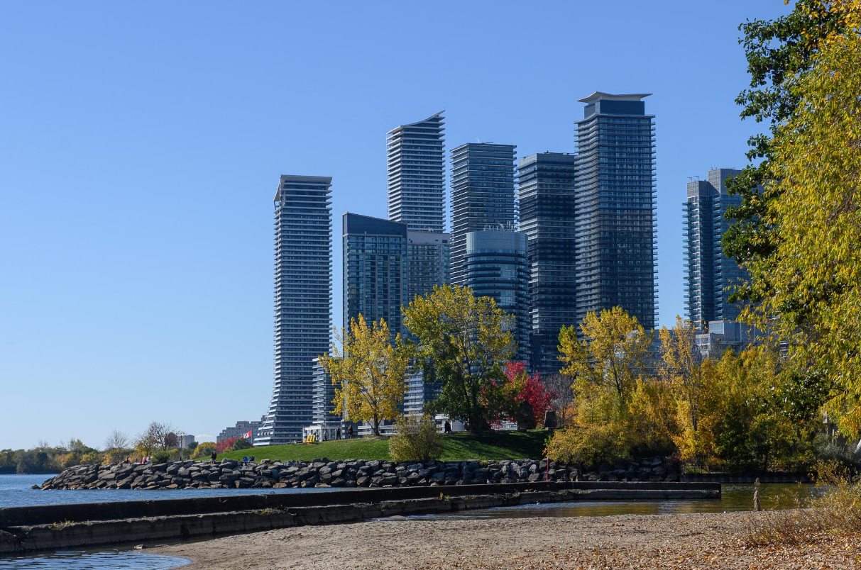 City skyline with tall modern skyscrapers, trees with autumn leaves, and a body of water in the foreground on a clear day.