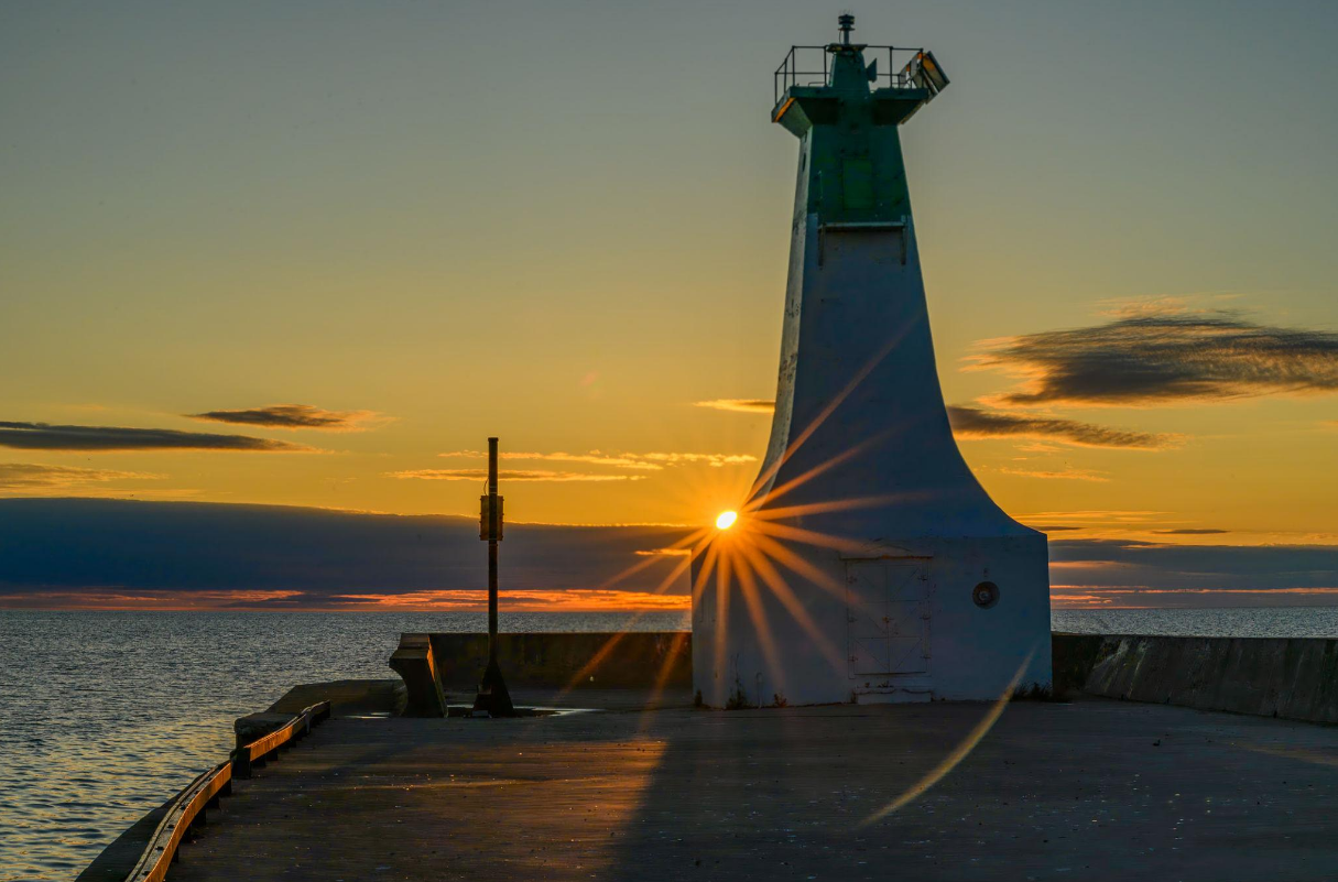 A lighthouse on a pier at sunset with the sun shining through the lighthouse, over a body of water.