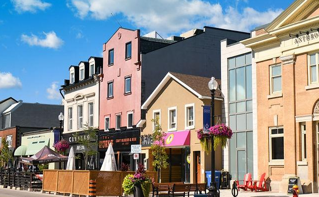 Colorful storefronts and buildings along a street with outdoor seating, umbrellas, flowering planters, and a clear blue sky.