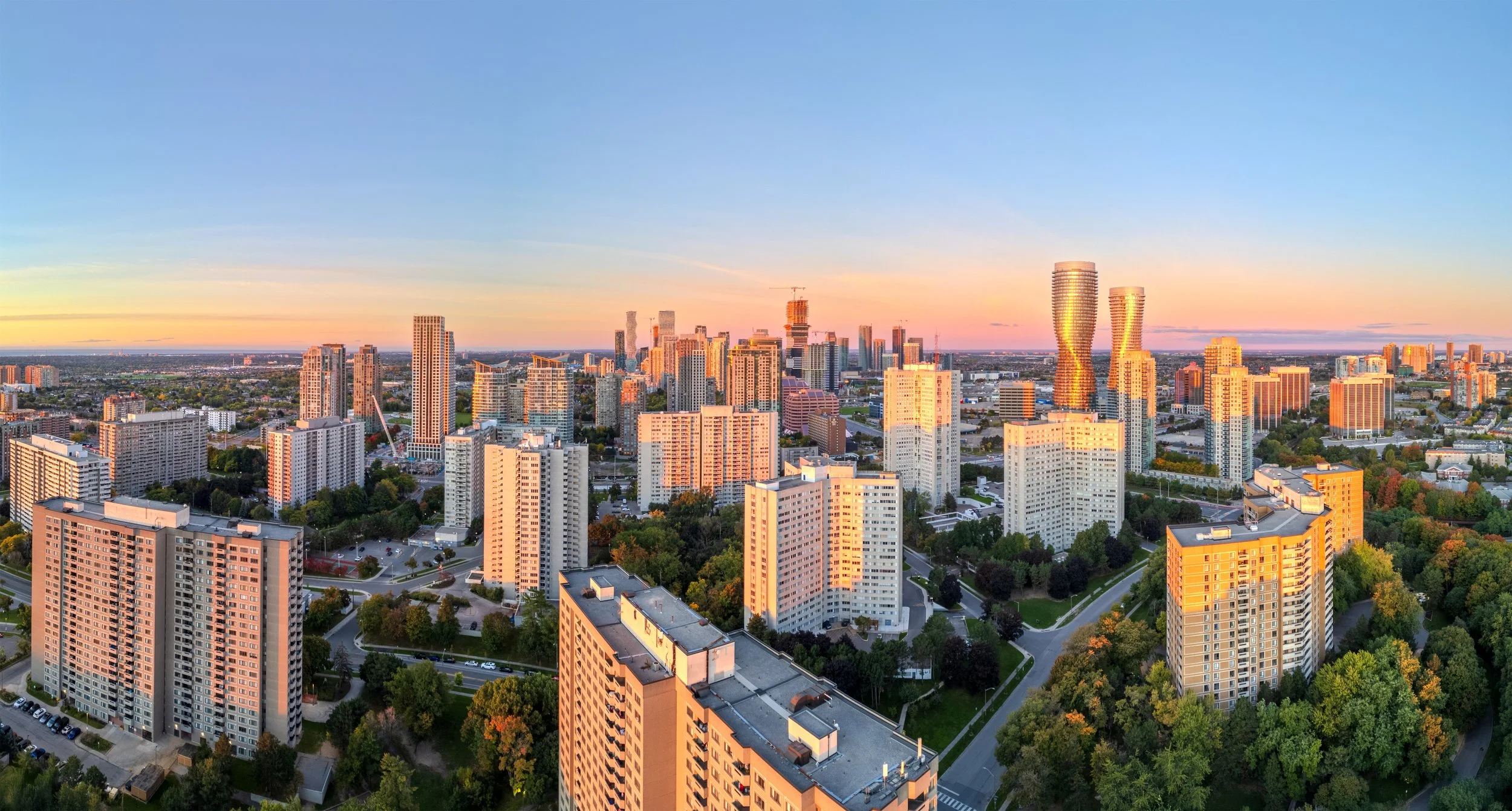 Aerial view of a city skyline during sunset, featuring tall modern skyscrapers, residential buildings, green trees, and a clear sky with the setting sun casting a warm glow.